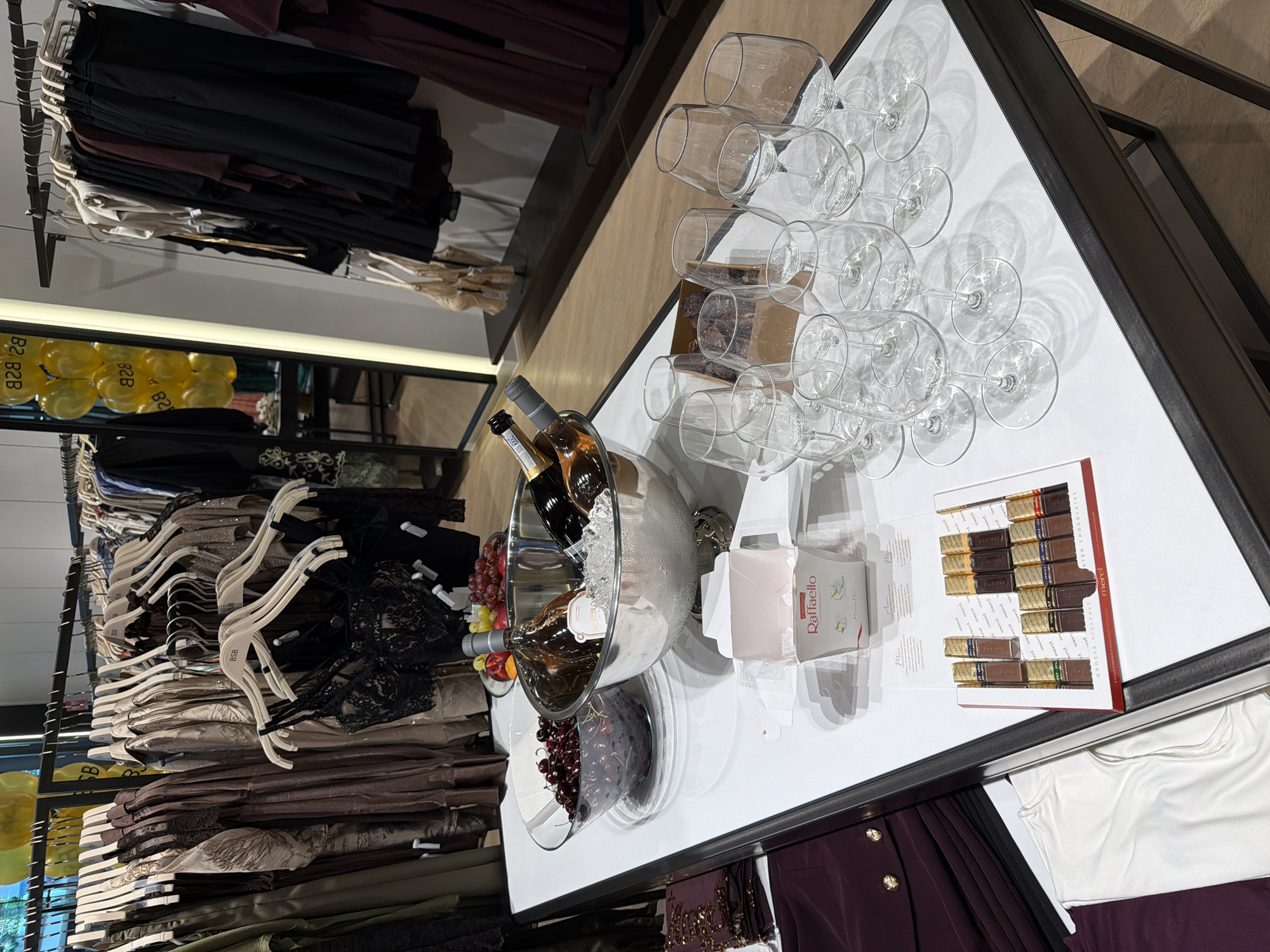 The image depicts a retail display in Burgas, Bulgaria, featuring clothing on racks in the background and a table in the foreground with various items. The table is set with several wine glasses, two bottles of wine chilling in an ice bucket, a bowl of cherries, a bowl of grapes and peaches, and two boxes of chocolates, one branded "Raffaello" and the other "merci". Partially visible in the lower left of the image are folded clothing items. The lighting suggests an indoor retail environment, possibly a boutique or department store. The overall scene suggests a promotional event or product display.