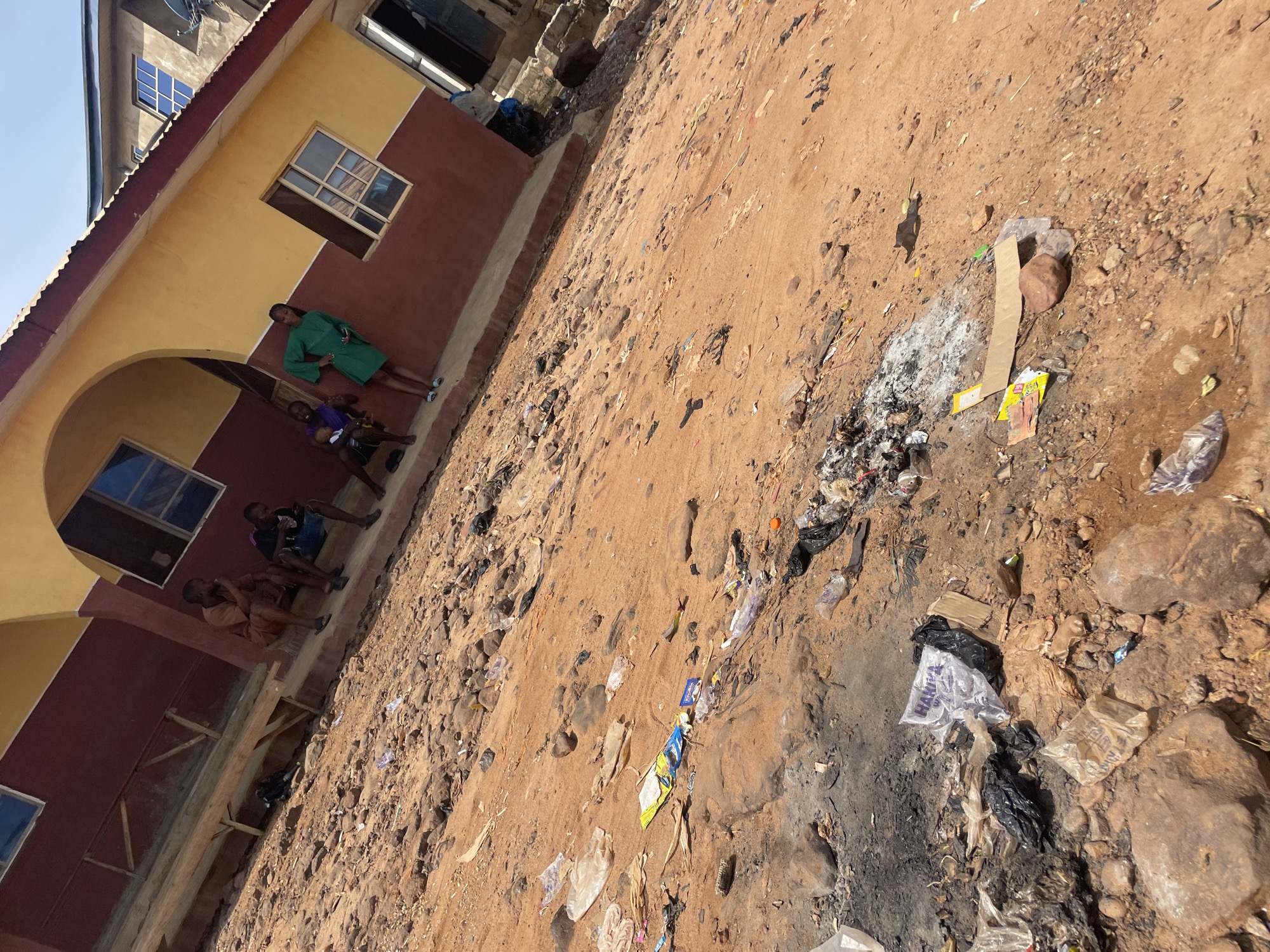 The image depicts an outdoor scene in Lokoja, Nigeria, characterized by a dry, reddish-brown dirt ground littered with various debris, including plastic bags, wrappers, and what appears to be burnt material. A building with a yellow and maroon facade stands in the background, featuring several windows and an arched entryway. A wooden bench is positioned against the wall.
Several individuals are visible:
* On the steps in front of the building, three young males are seated, with one appearing to be holding a mobile phone.
* Further to the right, a young female in a green dress stands near the building.
* Next to her, another individual, possibly a child, is seated, being held by someone partially visible.
* A fourth male is seated on the ground, to the left of the building's entryway.
The overall environment suggests an area that may be a public space, possibly a school or community building, given the presence of children and the seating. The significant amount of litter on the ground indicates a lack of waste management or a heavily trafficked area. The time of day appears to be daytime, with bright sunlight casting shadows.