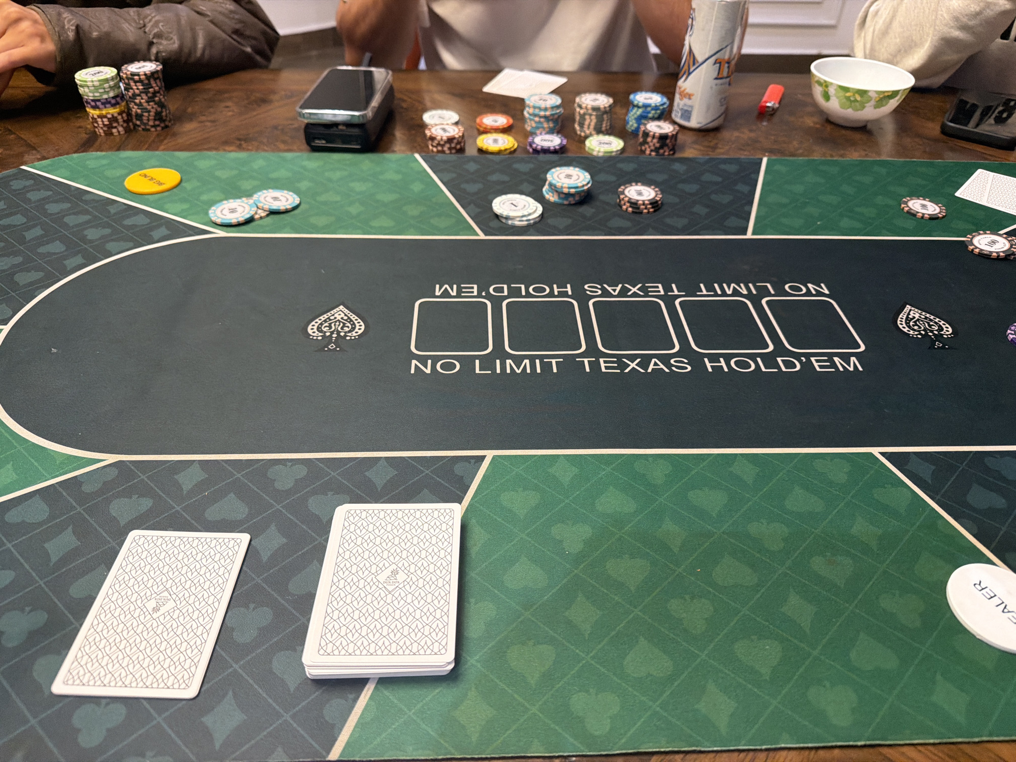An overhead, eye-level shot captures a poker game in progress on a green and dark green patterned felt table. The table is adorned with playing card suit symbols. In the foreground, two stacks of white playing cards, face down, sit on the green portion of the table. A white chip marked "DEALER" is visible to the right. The dark green portion of the table features the text "NO LIMIT TEXAS HOLD'EM" and four rectangular spaces for cards.
Scattered on the table are various poker chips in different colors, including white, blue, red, yellow, and green, some stacked and some loose. A single orange poker chip with the text "BIG BLIND" is also present. A cell phone, a red lighter, and a can of what appears to be beer are on the wooden surface behind the poker table. Two individuals, partially visible in the background, are seated at the table, suggesting they are participants in the game. The overall environment appears to be an indoor setting, possibly a home or a dedicated game room. The image does not provide any information that would allow for its location to be specified within Xuân Trường - Đà Lạt, Vietnam.