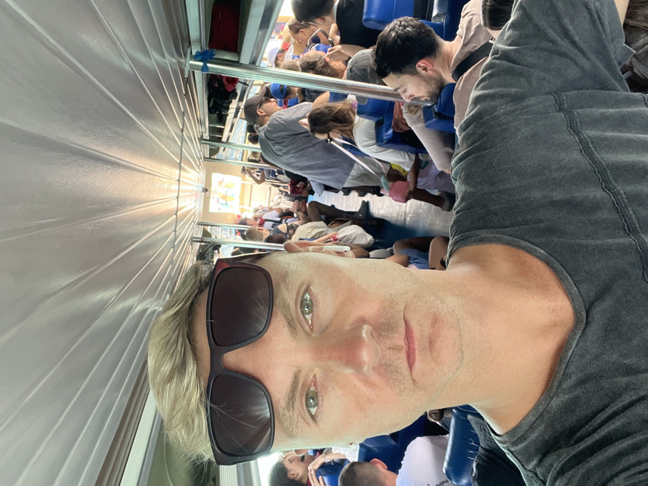 An interior view of a passenger vessel, likely a fast boat or ferry, is depicted, specifically within the harbor area of Toyapakeh, Indonesia. The central subject, a Caucasian male with short blonde hair and light green eyes, approximately 30-50 years old, occupies the foreground, taking a selfie. He wears a dark grey V-neck t-shirt and dark-rimmed sunglasses pushed onto his forehead, with a white earbud in his right ear. His expression is neutral.
The vessel's interior is brightly lit, featuring a light-colored, ridged ceiling and rows of blue passenger seating arranged longitudinally. Numerous passengers of varying ages and ethnicities occupy these seats, indicating a full capacity. In the middle ground, a man in a grey shirt and cap stands while others are seated. A child is visible holding a pink object. Luggage or bags are stored on overhead racks towards the aft section of the vessel. Bright daylight from an open section at the far end of the boat suggests the vessel is either docked or in transit in open waters, consistent with a port departure or arrival.