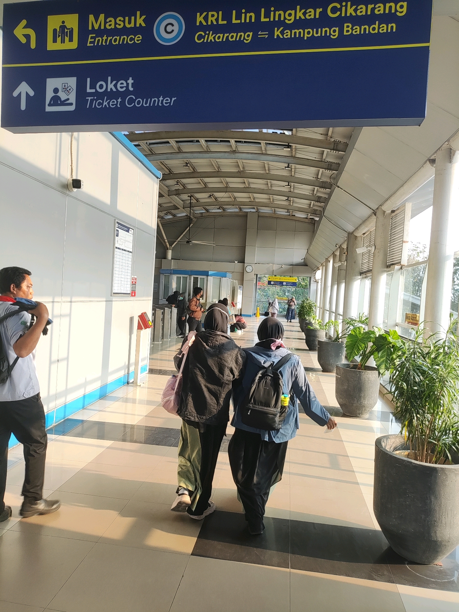 The image captures an indoor walkway, likely at a transportation hub in East Jakarta, Indonesia, characterized by its clean, modern design with white walls and a tiled floor. A prominent blue sign with yellow lettering indicates directions: "Masuk Entrance" with an arrow pointing left, and "Loket Ticket Counter" with an arrow pointing up. Another sign, partially visible, reads "KRL Lin Lingkar Cikarang" and "Cikarang Kampung Bandan".
Several individuals are present in the scene. In the foreground, a person wearing a light blue shirt and dark pants walks away from the camera, carrying a bag over their shoulder. To their right, two individuals, one in a black hijab and jacket, and another in a dark hijab and jacket with a backpack, are walking towards the background. Further in the background, more people are visible, some queuing at what appear to be ticket gates or turnstiles.
The environment is well-lit, with natural light streaming in from the right side, casting long shadows across the walkway. Large potted plants are strategically placed along the right side of the walkway, adding a touch of greenery. A white wall with blue trim runs along the left side of the walkway, and a security camera is mounted on it. The ceiling features a metal structure with translucent panels, suggesting it's a covered walkway. The overall impression is one of an orderly and functional public space.