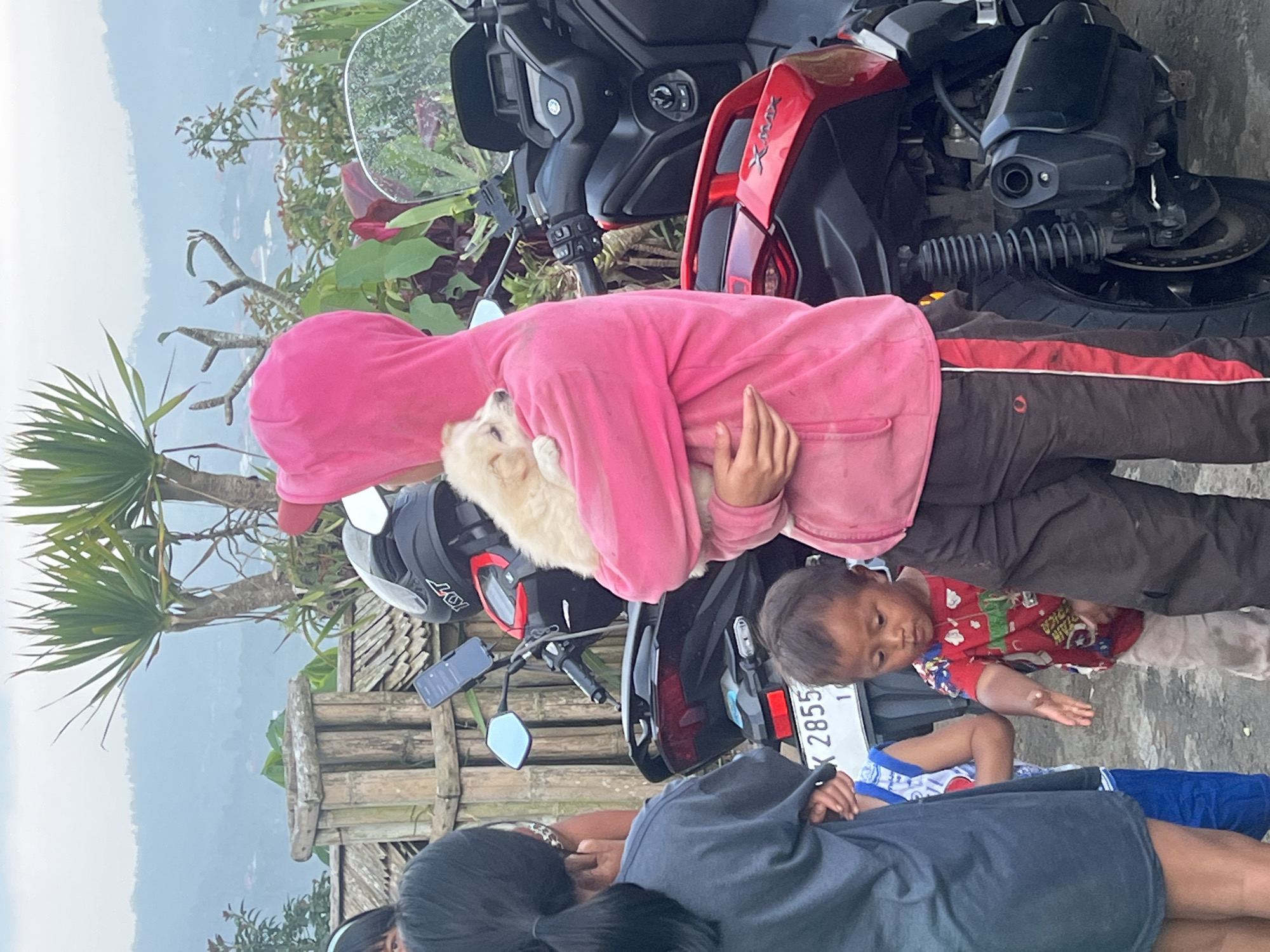 The image depicts a group of people and a dog next to a parked motorcycle in Kintamani, Indonesia. A person in a pink hooded jacket is holding a small, fluffy dog. A young child in a red shirt stands nearby. Another person, wearing a dark blue shirt, is also present. The motorcycle is a dark-colored model with red accents. A license plate is visible on the motorcycle. The surrounding environment includes trees and a bamboo fence, with a landscape view of the Kintamani region in the background.