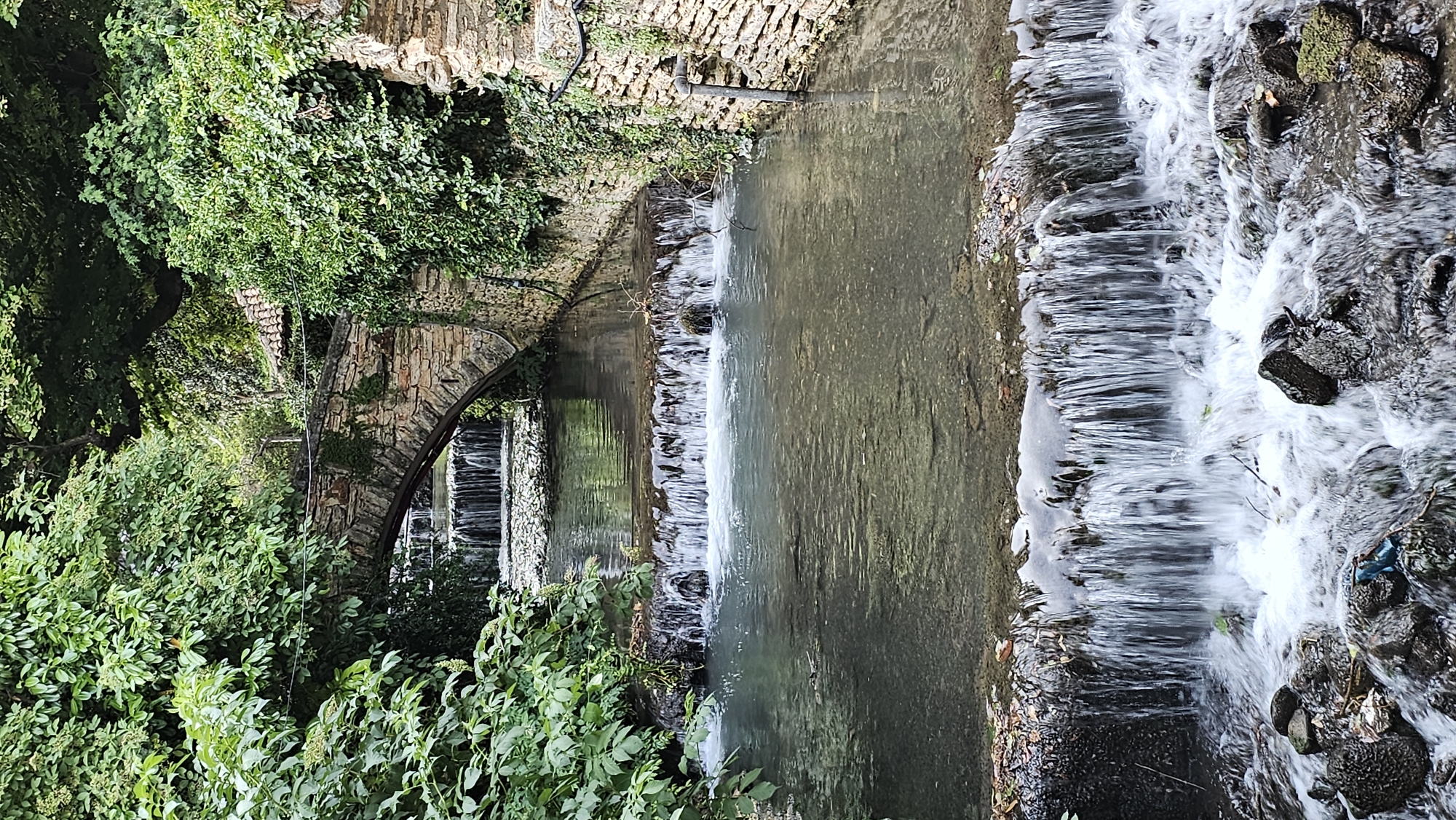The image depicts a scenic view of a small waterfall cascading over rocks and stone structures in Balchik, Bulgaria. The scene is dominated by lush green foliage encroaching on a weathered stone archway, suggesting an old, possibly historical, structure. The water is clear, with white foam indicating the motion of the falls. The overall impression is one of a tranquil, natural setting integrated with man-made elements. The time of day appears to be daytime, with diffused lighting that doesn't cast strong shadows, hinting at an overcast or lightly cloudy sky. There is no visible text in the image. The composition, with the water flowing from the upper right to the lower left, creates a dynamic visual flow. The presence of moss and a slight unevenness in the stone suggest age and exposure to the elements.