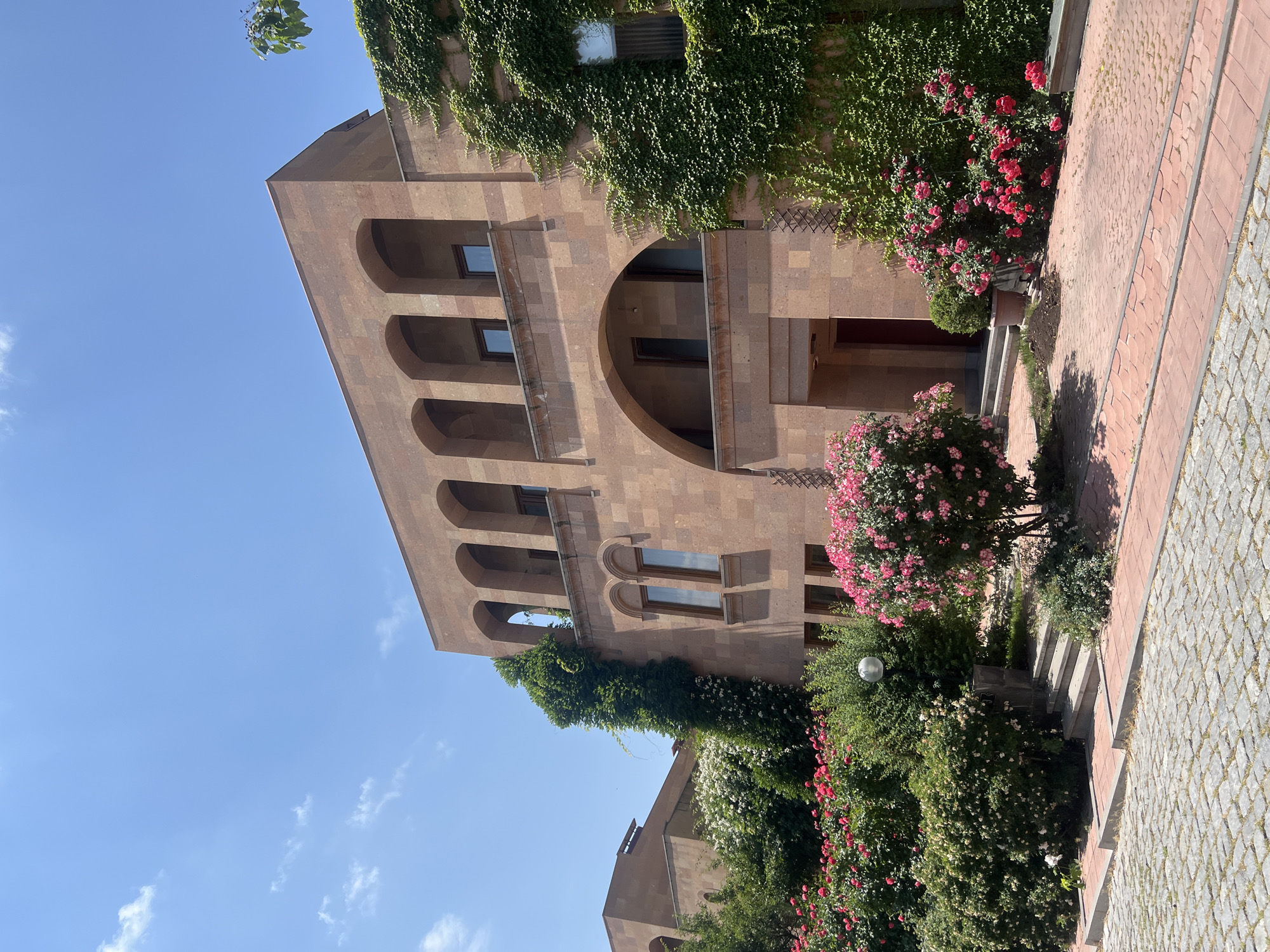 The image depicts the exterior of a building, likely in Yerevan, Armenia, characterized by its distinctive architecture and lush surroundings. The building is constructed from a warm-toned stone, possibly tuff, common in Armenian architecture. It features a series of arched windows and balconies on the upper floors, and a larger arched entrance. Ivy climbs prominently up the side of the building, adding a touch of natural greenery to the stone facade.
In the foreground, vibrant rose bushes in full bloom, displaying shades of pink and red, are a dominant feature. These flowers are arranged in a well-maintained garden setting, with interspersed green bushes and some smaller, manicured shrubs. The ground is paved with cobblestones in the immediate foreground, transitioning to a red-tiled path that leads towards the building's entrance. Steps are visible, providing access to the building. A single spherical light fixture is visible amongst the greenery.
The sky above is a clear, bright blue, with a few scattered white clouds, suggesting a sunny day. The overall impression is one of a serene and picturesque environment, likely a public space or a well-kept private residence, with a strong integration of natural elements with architectural design. The style of the building, particularly the extensive use of arched openings and the stone material, is highly characteristic of buildings found in Yerevan.