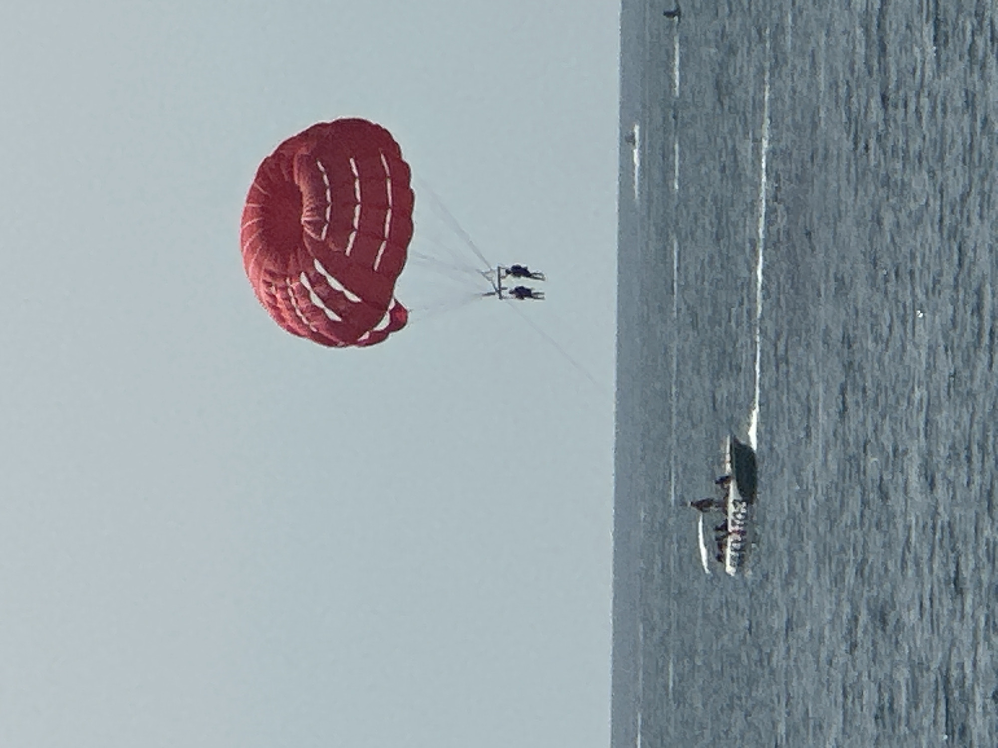 In the waters off the coast of Budva, Montenegro, two individuals are parasailing beneath a vibrant red parachute against a pale blue sky. A speed boat, labeled "PREDATOR-3," with several passengers aboard, navigates the choppy, dark blue sea in the foreground. The horizon line separates the sea from the sky, with no visible landmass. The image captures a moment of recreational activity on a clear day.
