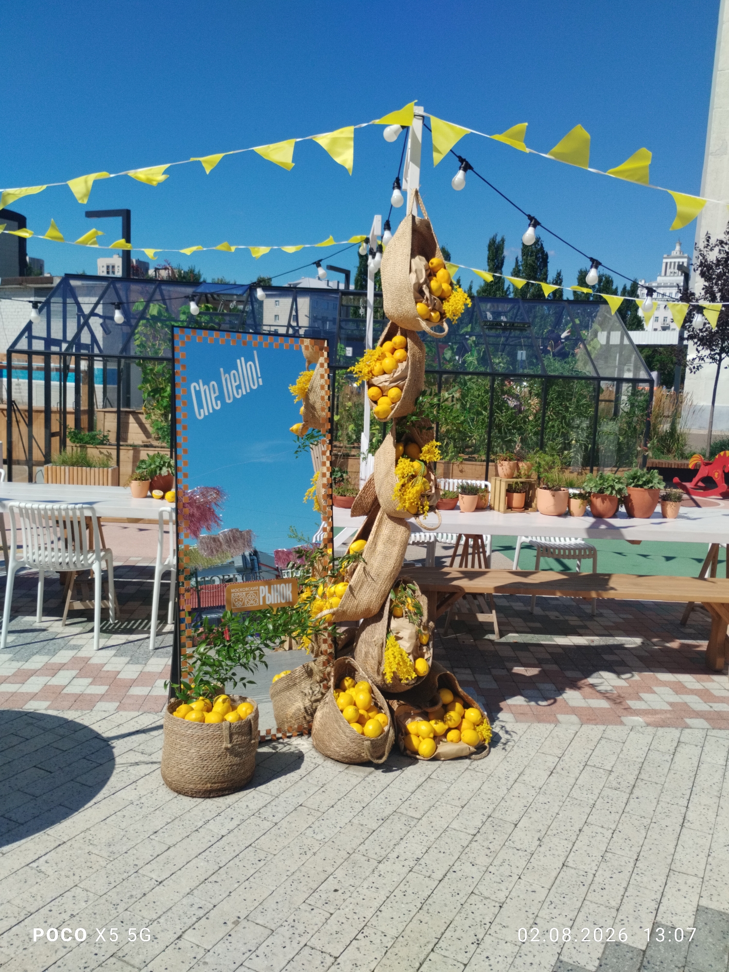 This image captures an outdoor market or cafe setting under a bright blue sky. Yellow triangular flags are strung across the top of the frame, and string lights with exposed bulbs are also visible. A prominent feature is a decorative wooden structure wrapped in burlap, adorned with woven bags filled with lemons and yellow flowers. A large rectangular mirror with the text "Che bello!" on it stands in front of the structure. In the background, there are greenhouses, tables with chairs, and a wooden bench. The ground is paved with light-colored tiles. The lower right corner of the image displays the text "02.08.2026 13:07". The lower left corner shows "POCO X5 5G".
