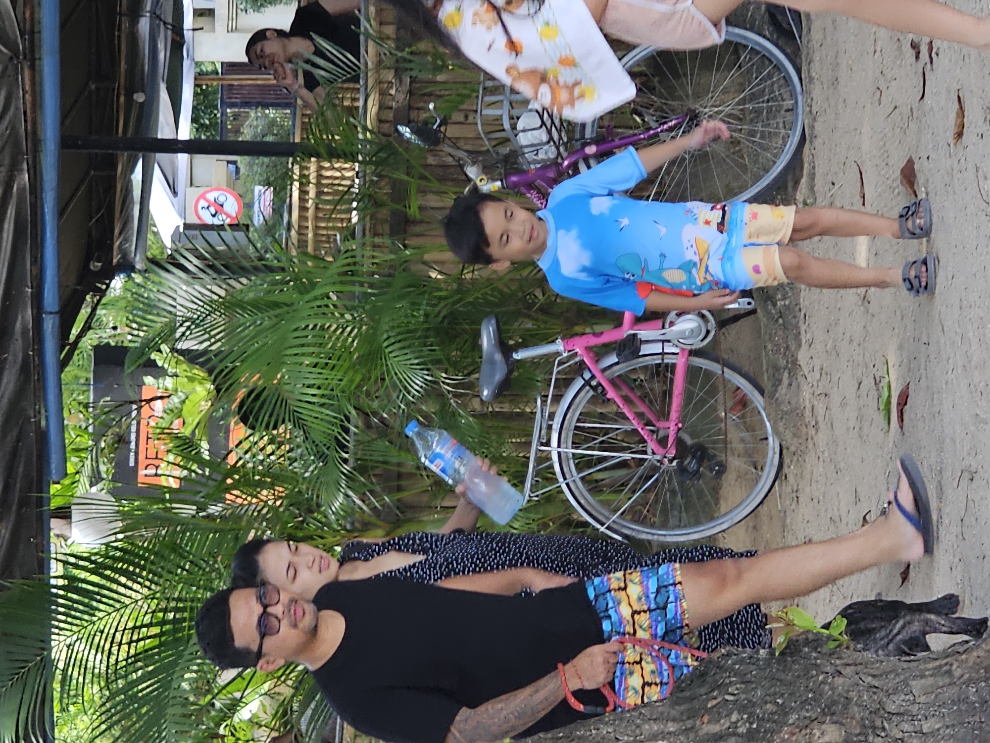 The image shows a partly shaded outdoor setting, likely in Denpasar, Indonesia. In the foreground, a man wearing a black t-shirt and patterned shorts stands partially behind a tree, with a woman in a polka dot dress leaning on him. A water bottle is near them.
Further back and to the right, a young boy in a blue shirt and beach-themed shorts stands near a pink bicycle. Behind the boy, there's another bicycle with a purple frame and a fair-skinned person wearing white and pink clothing. In the upper left corner, a man can be seen in a raised shaded area. There is lush tropical vegetation throughout the shot. A street sign is visible. The ground is mostly sandy with some vegetation.