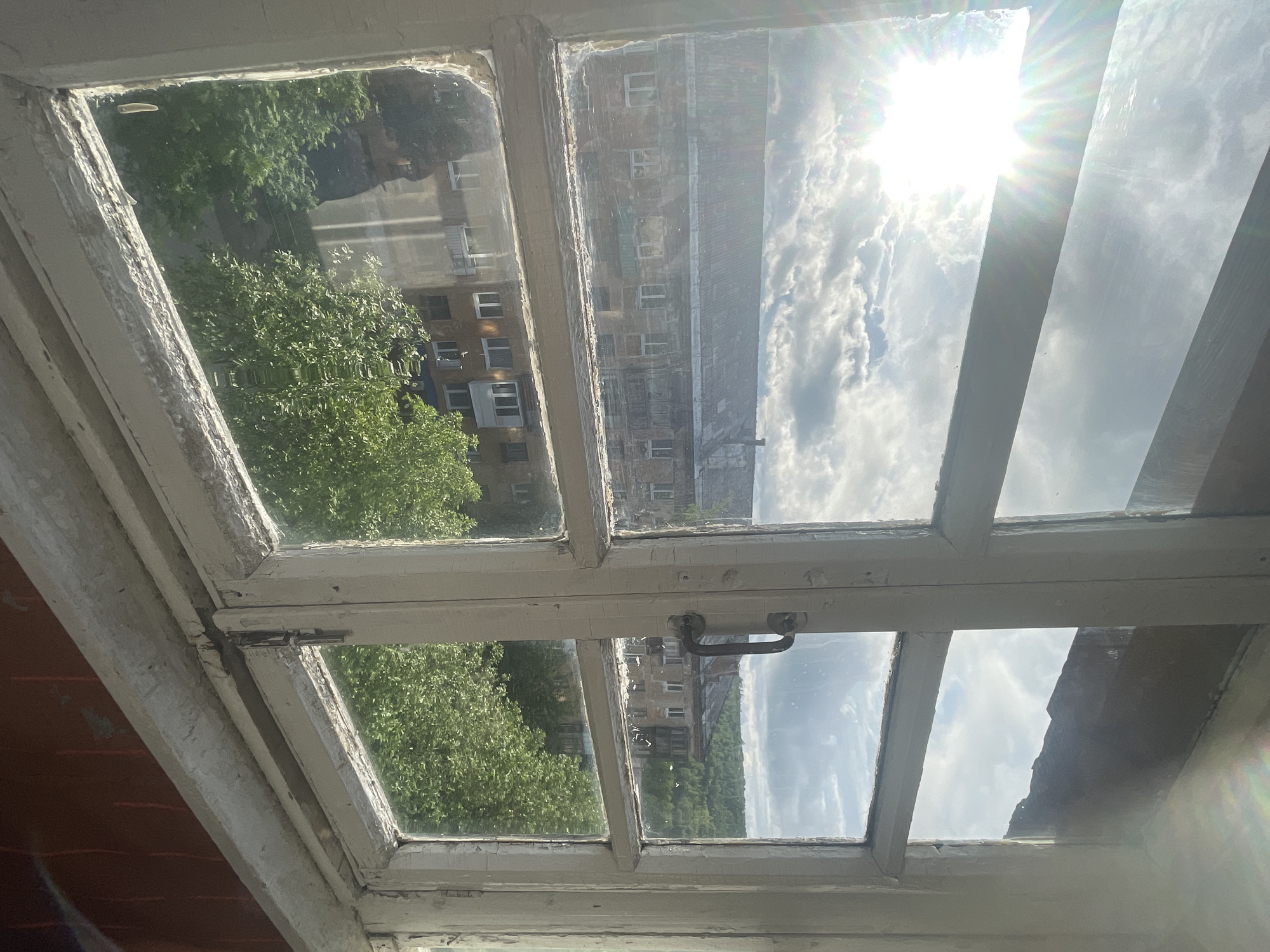 The image presents a view from an interior space looking outwards through an old, white-painted wooden window frame with multiple glass panes. The window frame exhibits significant wear, with extensive peeling paint and visible dirt or dust accumulation in crevices. A black metal handle is affixed to the central vertical mullion on the right side. The windowsill, also painted white, shows similar signs of aging and wear. A reddish-brown wall or surface is visible at the bottom right corner, partially obscured.
Outside the window, a bright, partly cloudy sky is visible. A prominent sunburst lens flare emanates from the upper-left pane, indicating direct sunlight. In the middle ground, old, multi-story residential buildings with dark, corrugated roofs are visible. These buildings appear to be constructed of brick or stucco, exhibiting a weathered and somewhat dilapidated appearance with numerous windows. Below these structures, dense green foliage from trees is prominent, suggesting a street-level or courtyard view. On the right side of the view, beyond the buildings and trees, a distant landscape of green, tree-covered hills or mountains can be observed under the sky. No individuals, explicit actions, or specific distinguishing objects are discernible within the frame. The scene depicts an aged urban or semi-urban residential environment, consistent with areas found in the city of Kizel, Russia, characterized by Soviet-era architecture and surrounding natural landscapes. The bright sun suggests a daytime setting, possibly morning or late afternoon.