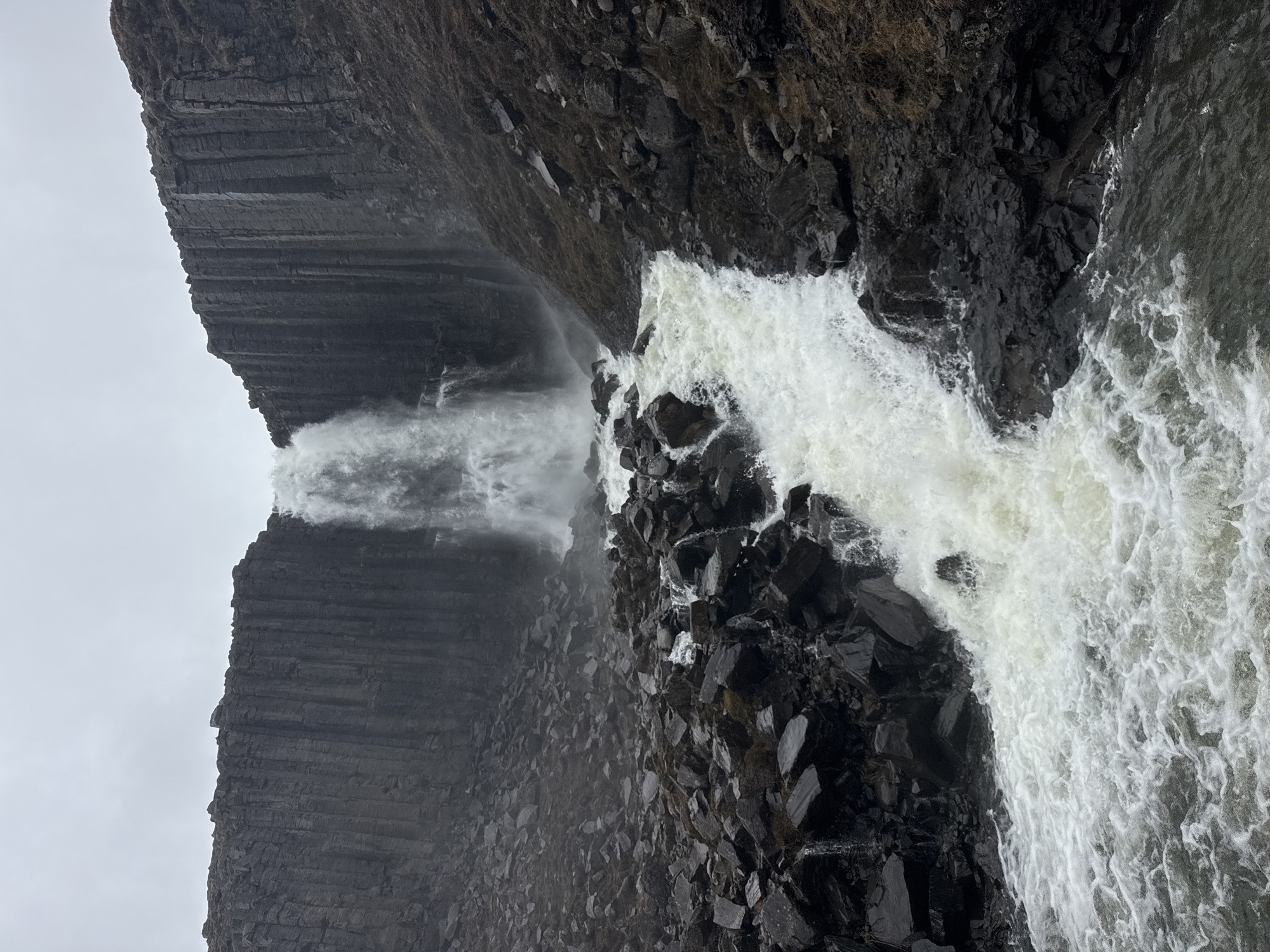 The image, originally rotated 90 degrees clockwise, depicts a powerful waterfall cascading down a dark, rocky cliff face. A prominent feature on the left (when oriented correctly) is a massive cliff displaying distinct horizontal layers and vertical fractures, characteristic of columnar basalt. Sparse, dry vegetation can be seen clinging to the upper sections of this rugged, dark rock formation.
The main body of the waterfall plunges forcefully, creating a wide, frothy, white torrent as it crashes onto a chaotic collection of large, jagged dark rocks at the base of the cliff. To the left of the primary cascade, a smaller, thinner stream of water also descends, partially obscured by the spray and mist generated by the main falls. The turbulent water below is churned into white foam, eventually flowing into a darker body of water, likely a river or pool.
The sky is a uniform, bright overcast grey, indicating a cloudy daytime setting with diffused light. No people, man-made objects, or visible text are present in the scene. The dramatic geological features, particularly the columnar basalt and powerful waterfall, are suggestive of an Icelandic landscape, as hinted by the provided context.