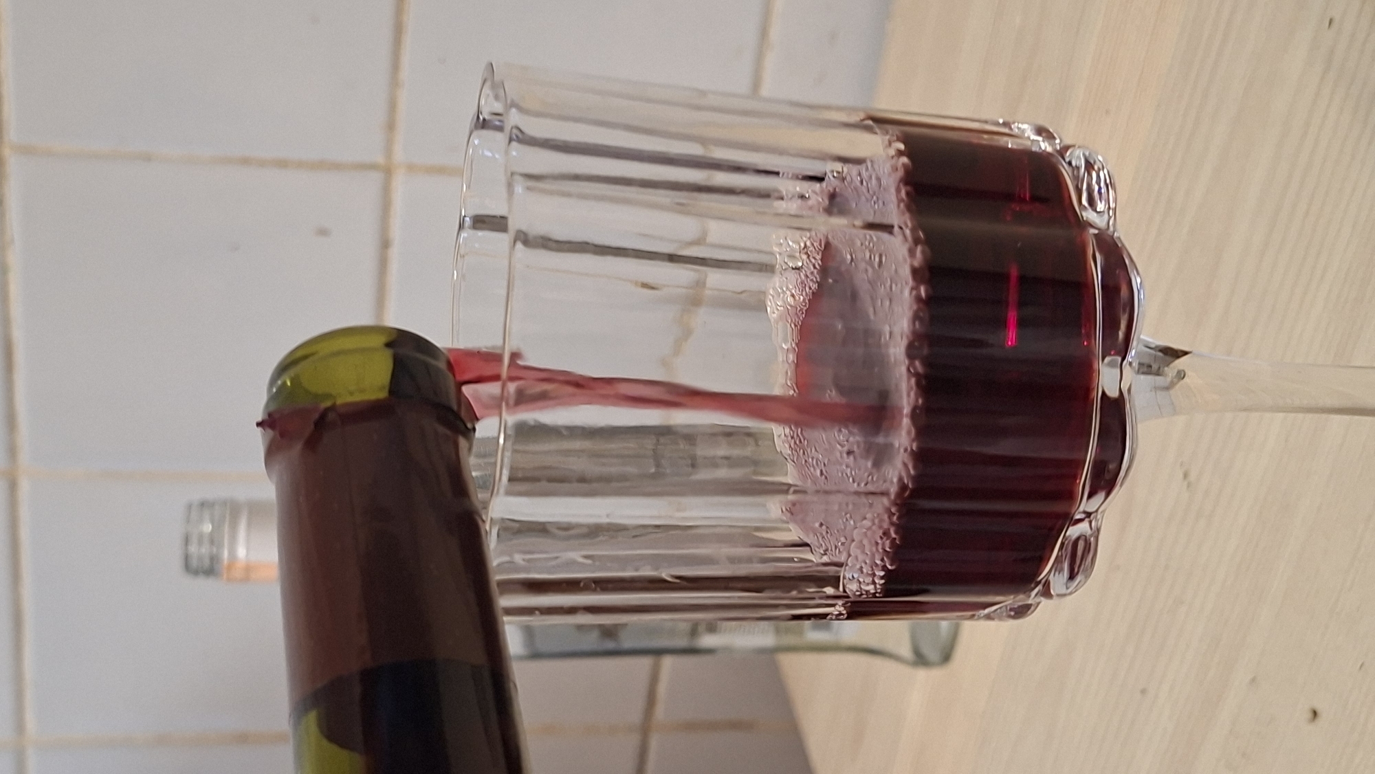 A close-up shot shows red wine being poured from a dark green bottle into a clear glass with vertical ridges. The wine is a deep red color and is creating a small amount of foam as it fills the glass. In the blurred background, another bottle with a silver cap is visible, and a tiled wall can be seen. The scene is set on a light-colored wooden surface. The location is likely a domestic setting in Забалоцкі сельскі Савет, Belarus.