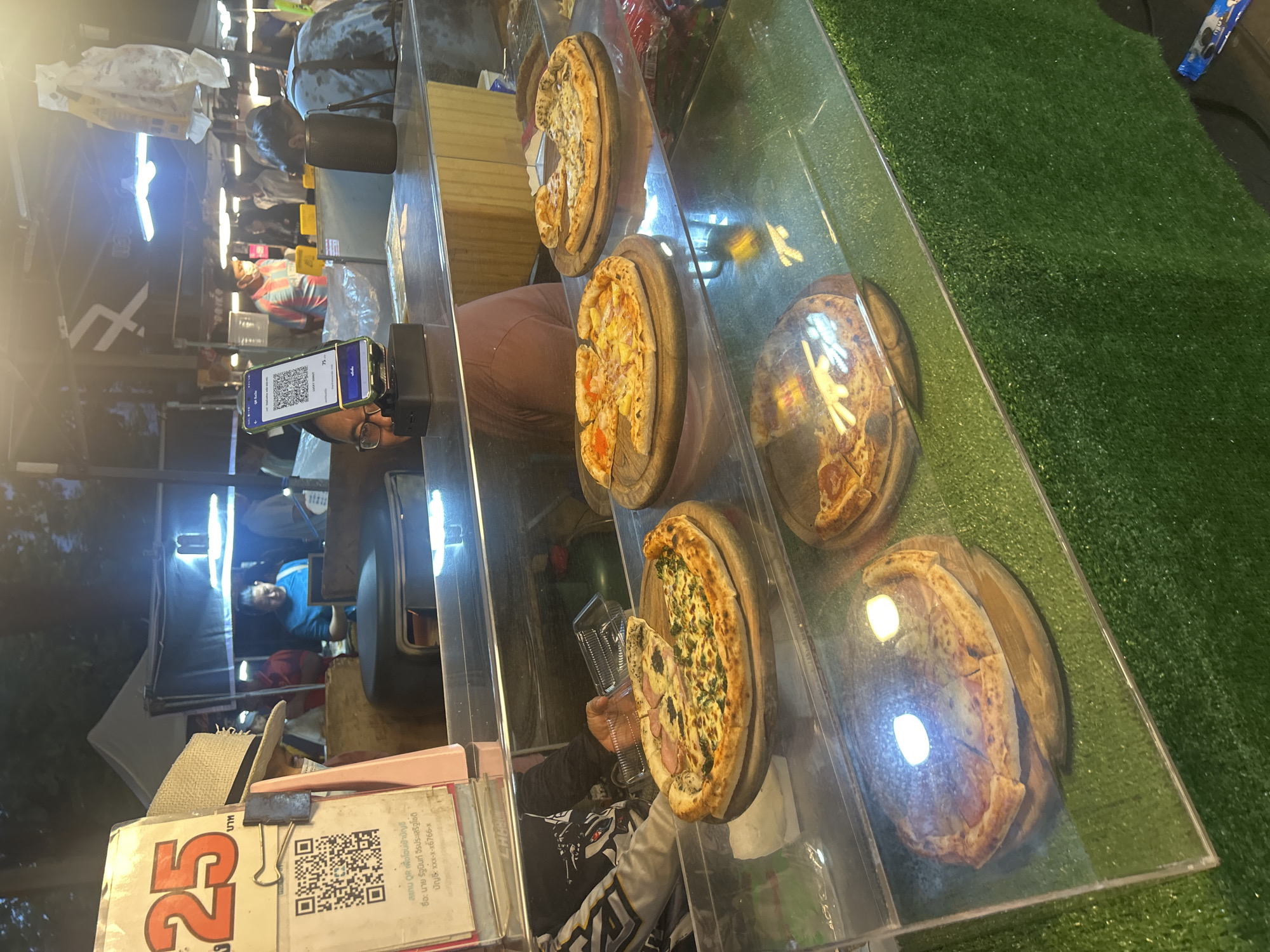 In Tha Khlong Town Municipality, Thailand, a street food vendor is selling pizzas from a stall. The stall is set up with a glass display case showcasing several pizzas on wooden boards. In the foreground, a large pizza with what appears to be spinach and ham is displayed. Behind it, other pizzas with various toppings are visible. A sign with the number "25" in red is prominently displayed on the left, possibly indicating a price. A QR code is also visible on the sign, suggesting a cashless payment option. A person wearing glasses and a dark shirt is partially visible behind the counter, interacting with the display. In the background, other vendors and customers can be seen, and tents suggest a bustling market environment. The overall setting is a nighttime market scene with artificial lighting illuminating the food stall.