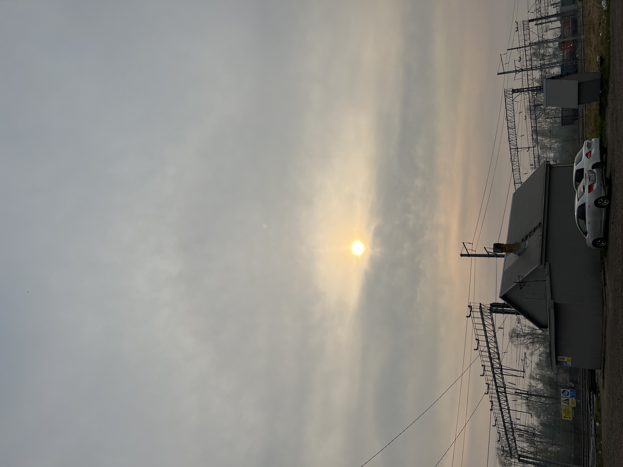 The image shows an exterior scene in an urban or industrial area. The sky is overcast with the sun visible through the haze. In the foreground, there is a gray, possibly metallic building with a peaked roof. Parked next to the building are three white cars. Overhead power lines and metal structures, likely associated with a railway, are visible in the midground. The location appears to be near a railway yard with industrial buildings and signage visible in the background.