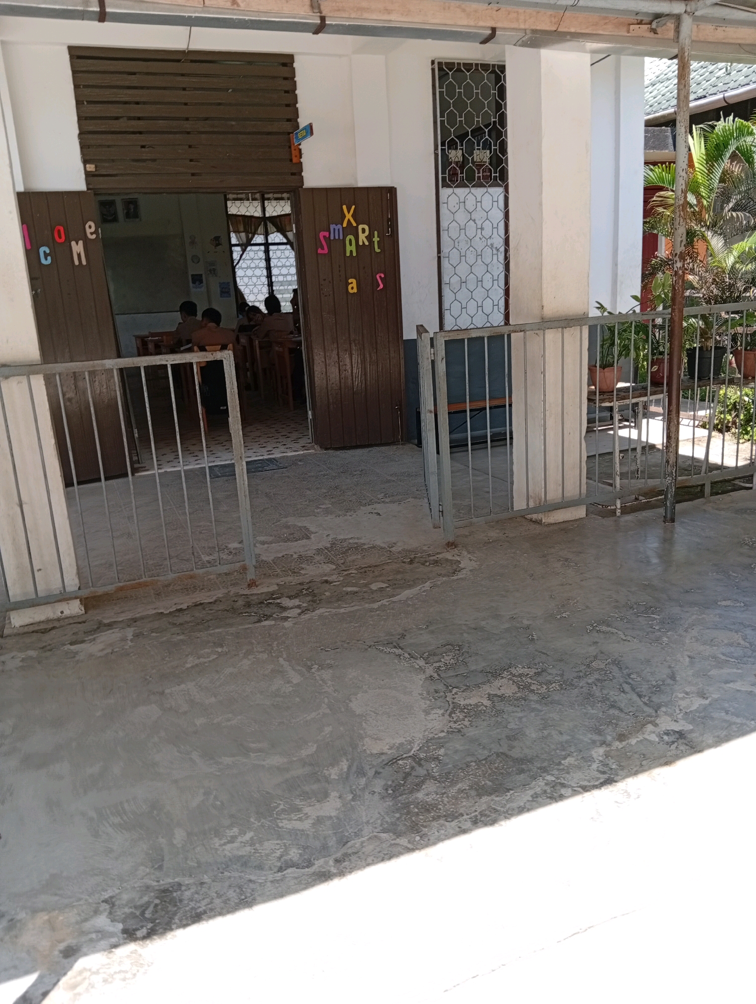 The image depicts the exterior of a building, likely a school or community center, in the City of Gunungsitoli, Indonesia. A metal fence with vertical bars stands in the foreground, partially obscuring a concrete walkway or patio. Through an open doorway, a classroom interior is visible, with several students in uniform seated at desks. Wooden doors flank the entrance, with colorful letters spelling "WELCOME" on the left door and "SMART KIDS" on the right door. The building features white walls and a tiled roof. Lush green plants in pots are arranged to the right of the entrance, suggesting an outdoor or semi-outdoor area. The overall scene suggests a bright, sunny day.