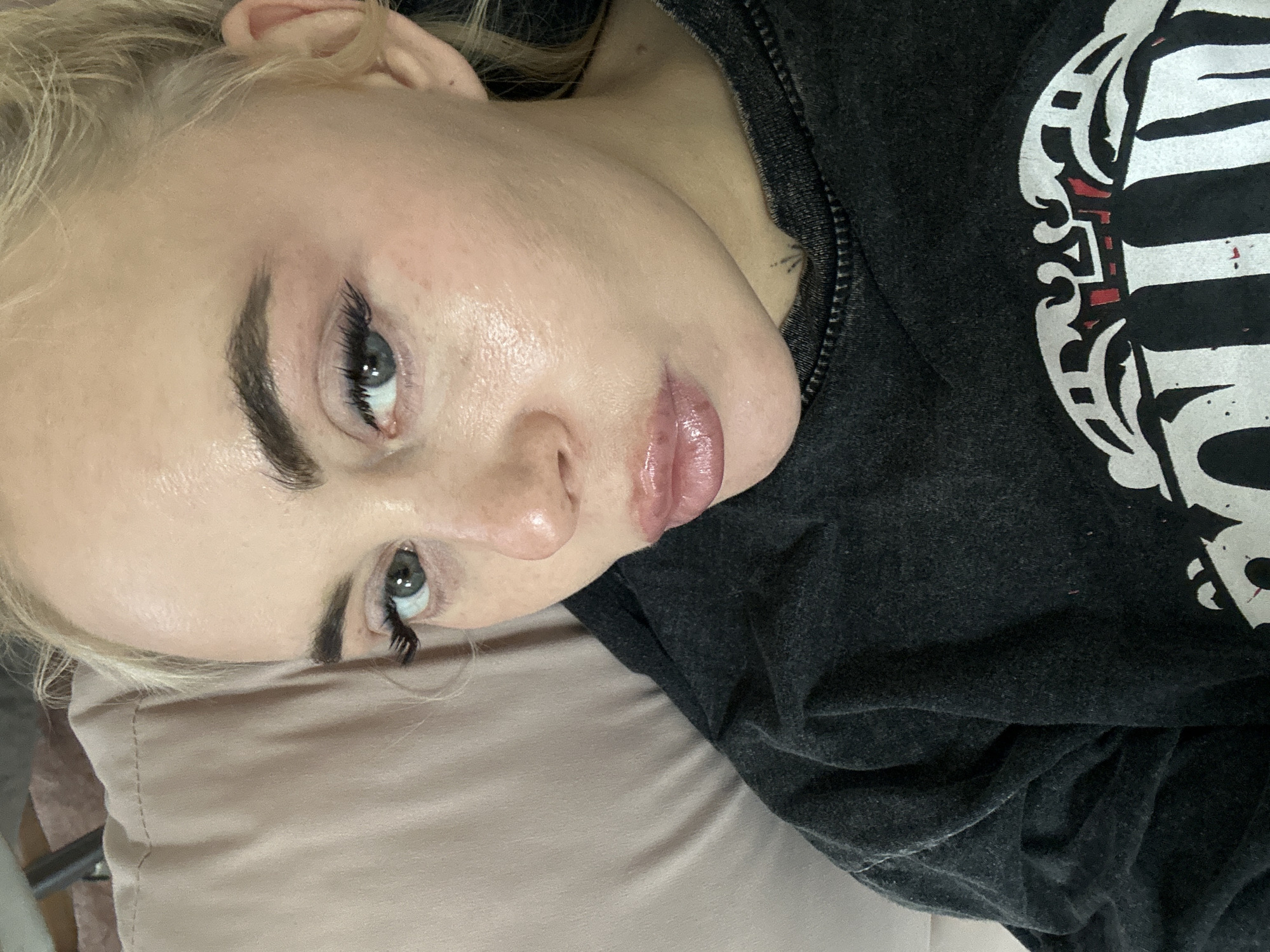 A close-up, tilted image captures a fair-skinned, blonde-haired person with visible makeup (eyebrows, eyelashes, lip gloss) lying on a light pink pillow. The person is wearing a dark, hooded sweatshirt with a white graphic design visible on the fabric. A small tattoo is located on the neck near the collarbone. The background is blurred. The image does not provide any information to establish a location in Высокий, Russia.