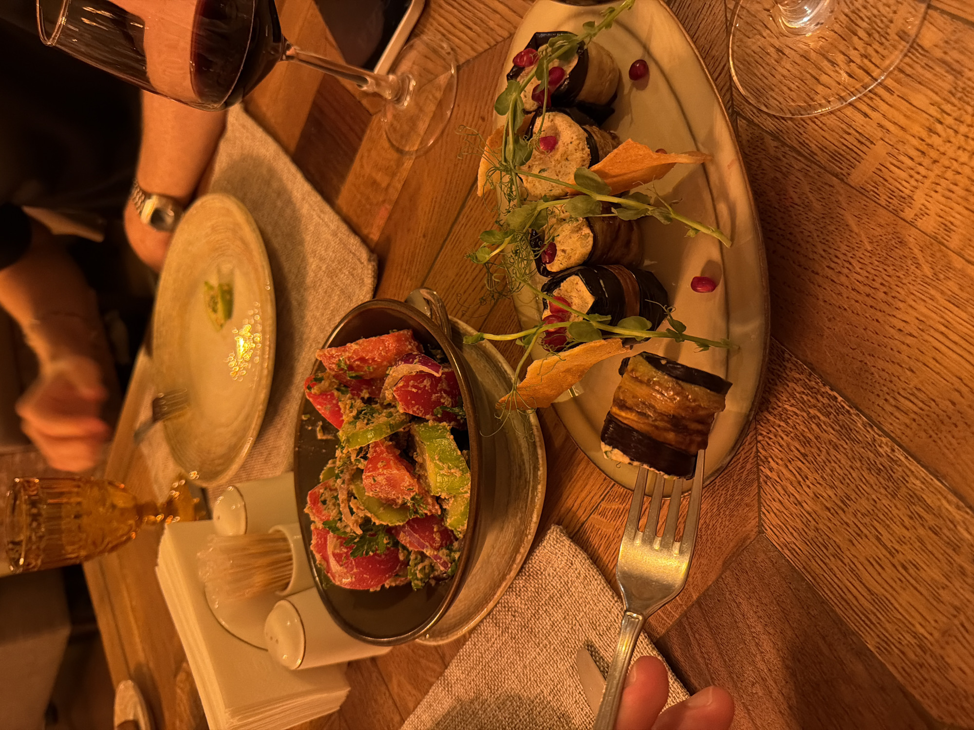 The image depicts an indoor dining scene at a restaurant in Tbilisi, Georgia, illuminated by warm, dim lighting. The focus is on a wooden table set for two individuals, showcasing traditional Georgian dishes.
In the foreground, a person's hand holds a silver fork, lifting a piece of Badrijani Nigvzit (eggplant roll with walnut paste) from an elongated, light-colored ceramic plate. This plate contains multiple servings of the eggplant rolls, each garnished with microgreens and scattered pomegranate seeds. Flat, orange-brown crisps are also present on this plate.
To the left of the eggplant rolls, a dark ceramic bowl holds a salad composed of chopped red tomatoes, green cucumbers, and herbs, mixed with a dressing and what appears to be ground walnuts.
In the mid-ground, a tall stemmed glass contains red wine, positioned near the right edge of the frame. Further back and to the right, a person's arm, wearing a dark shirt and a silver wristwatch, is visible. An almost empty light-colored plate with residual food and a fork rests on a beige linen placemat in front of this individual.
Other table items include a stack of white paper napkins, white ceramic salt and pepper shakers, and a container with toothpicks, all situated near an ornate, amber-colored glass containing a clear liquid, possibly water, in the background. The wooden table exhibits a pronounced grain pattern.