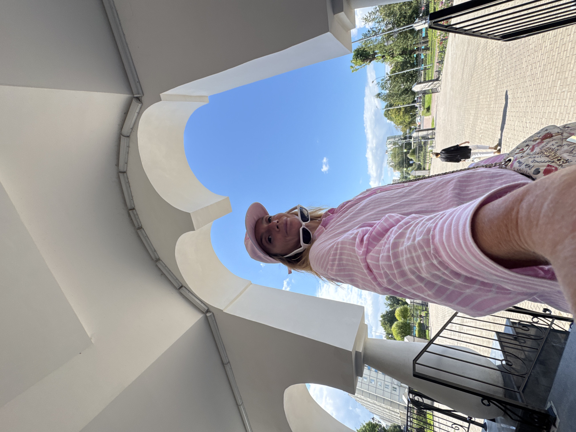 The image captures a selfie taken by a woman wearing a pink striped shirt, a pink baseball cap, and white-rimmed sunglasses. She is positioned in the foreground, looking at the camera. Behind her, an architectural structure with white arched elements frames a bright blue sky with scattered clouds. In the middle ground, a paved walkway leads to an open area with trees and buildings visible in the distance. Another person, wearing a black top and white pants, is walking away from the camera on the walkway. The overall setting appears to be an outdoor public space. The architectural style suggests a modern or renovated building, possibly in a park or a cultural complex. Without specific identifying landmarks, pinpointing the exact location within Saint Petersburg is not possible.