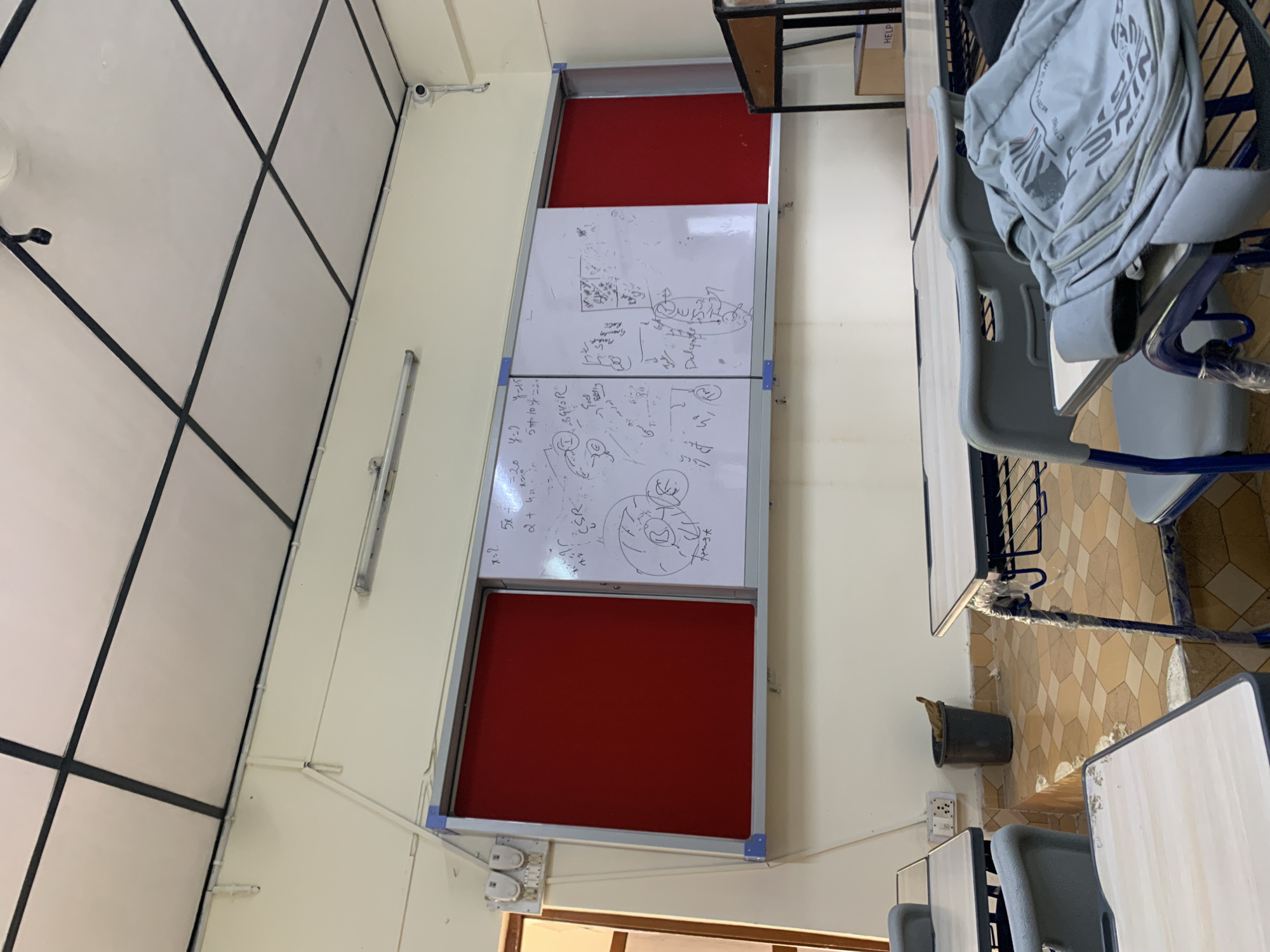 The image depicts a classroom setting in Dharamshala, India. A whiteboard, partially filled with handwritten notes and diagrams, is mounted on the wall. To the left of the whiteboard, a red bulletin board is visible. Several desks with chairs are arranged in the room. One chair has a gray backpack placed on it. In the background, a trash can is situated near a wall with electrical outlets. The room appears to be unoccupied at the time the photograph was taken. The overall environment suggests an academic or educational space.