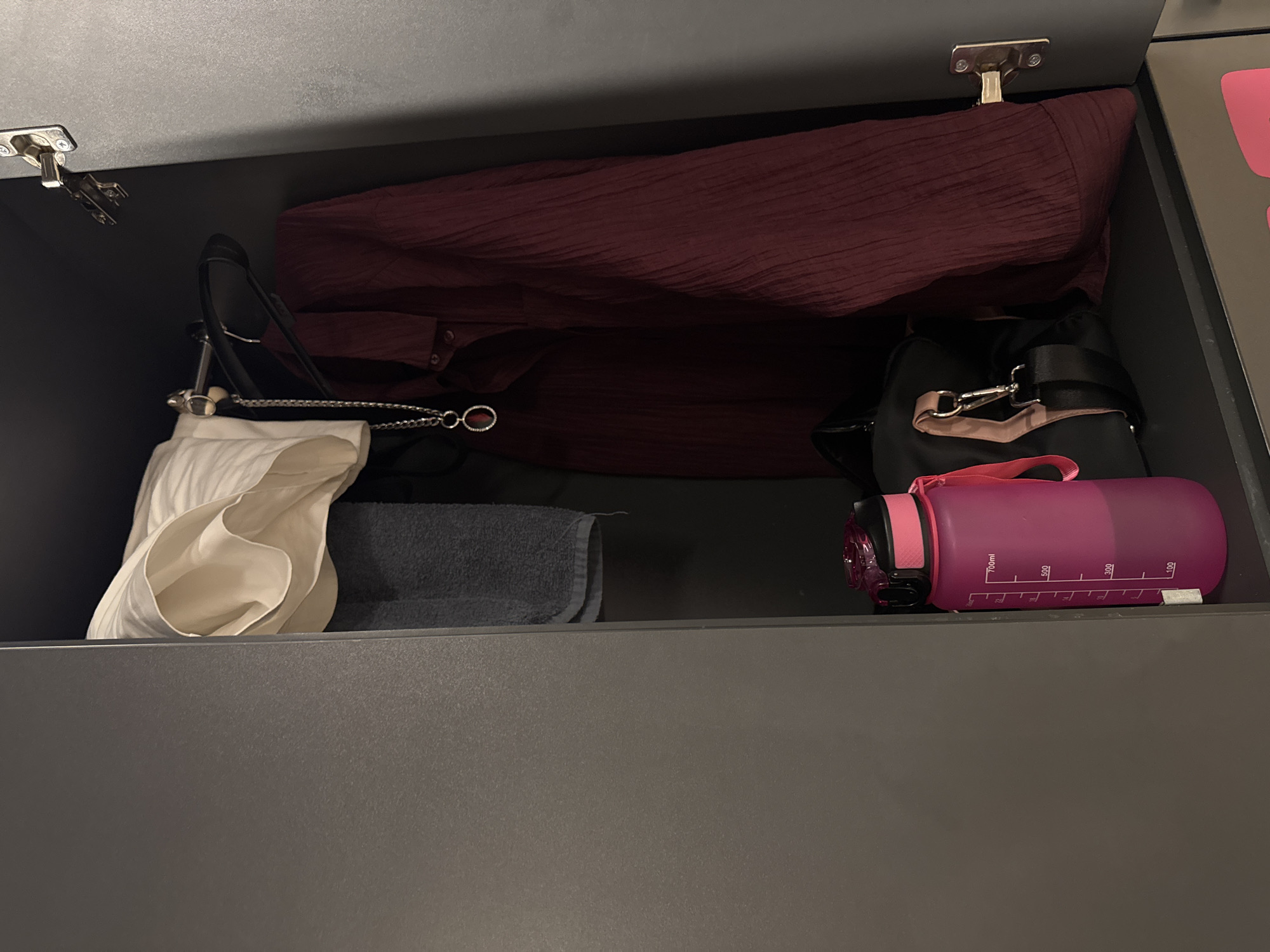 The image shows the inside of a dark gray cabinet. A large, textured, dark red garment is folded across the top. Below it, on the left side, is a white blouse or shirt folded with its sleeves draped over the side. To the right of the white garment, there is a dark gray folded towel. On the right side of the cabinet, a black bag with a pink strap is visible. Next to the black bag, a large pink water bottle with measurement markings is lying horizontally. The bottle has a black lid and a pink carrying strap attached. Visible text on the water bottle includes "700ml", "500", "300", and "100". The cabinet appears to be a storage space, possibly a drawer or a chest. The lighting suggests an indoor setting, with the light source illuminating the contents from above. There are no people or animals visible in the image. The overall scene is one of organized storage.