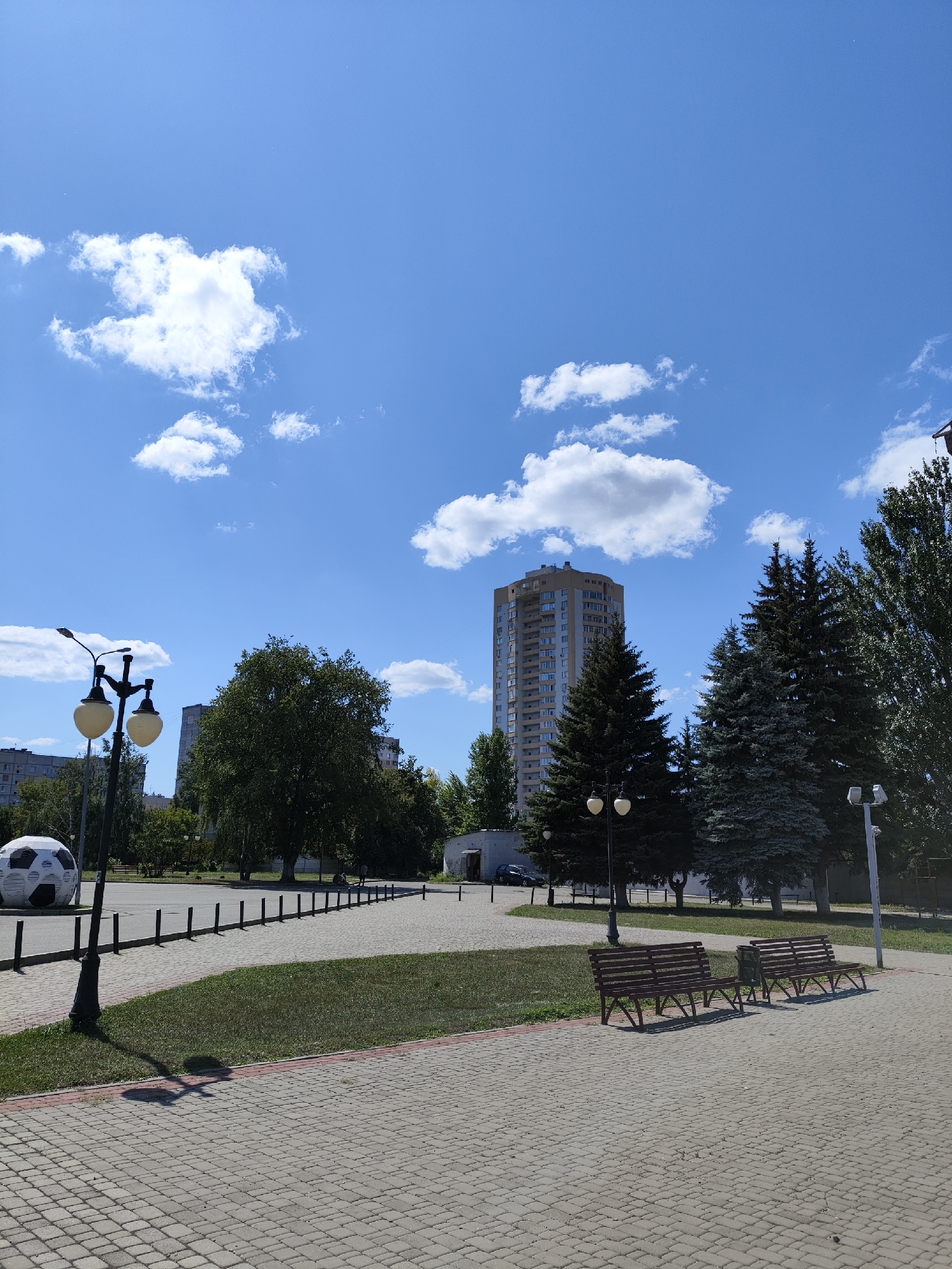The image depicts an outdoor scene with a clear blue sky dotted with fluffy white clouds. In the foreground, a paved walkway made of interlocking rectangular stones leads towards the right. Two brown wooden park benches are situated on a grassy area adjacent to the walkway.
Further into the scene, a large, black and white spherical sculpture resembling a soccer ball is visible on the left. A lamppost with two globes stands near the soccer ball sculpture. A row of short black posts runs along the edge of a paved area behind the walkway and grassy patch.
Dominating the middle ground are several mature evergreen trees and a large deciduous tree. In the background, a tall, multi-story apartment building with numerous windows stands out. Additional residential buildings are partially visible on the far left. The environment appears to be a public park or square within a city. Based on visual cues like the architecture and the presence of specific types of trees, this scene is likely located in the city of Kharkiv, Ukraine. The lighting suggests it is daytime, with sunlight casting shadows on the ground. No people are clearly visible in the frame.