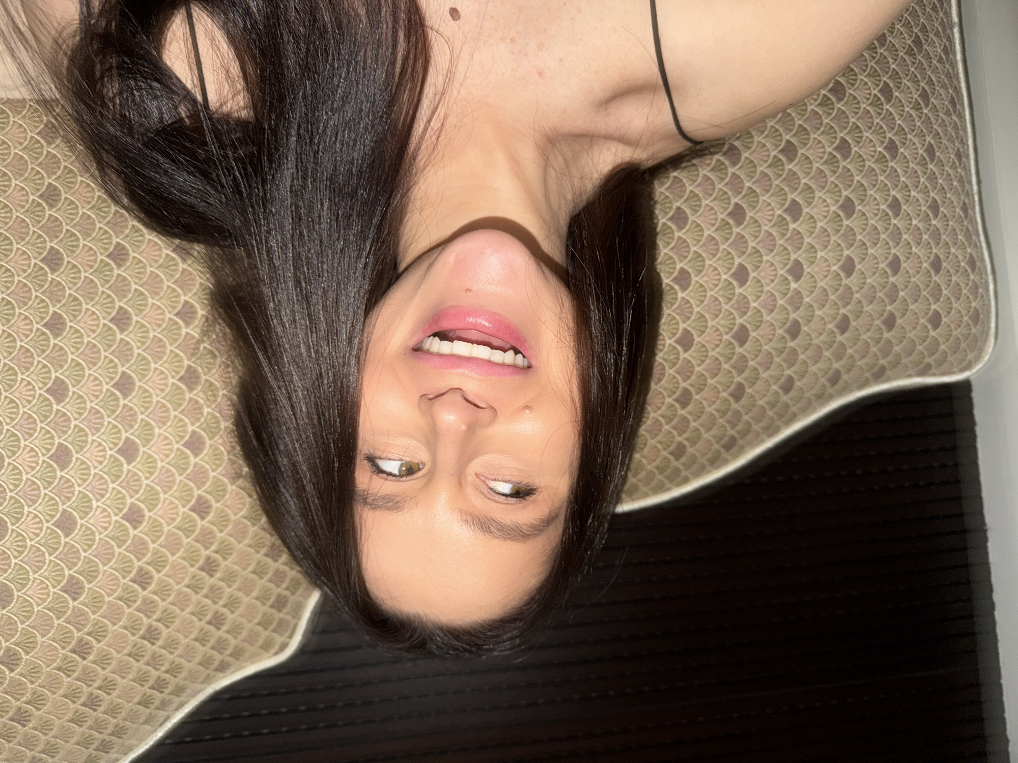 An upside-down close-up shot features a person, likely female, with long, straight dark hair framing her face. She is lying on her back, with her head closest to the bottom of the frame, giving the impression of being inverted. Her eyes are open and looking slightly upward and to the side, with a playful or lighthearted expression. Her mouth is open in a smile, revealing white teeth, and her lips appear to have a pink tint.
The person's neck and upper chest are visible, showing light skin tone with some freckles and a mole. A thin black strap, possibly from a piece of clothing, runs across her shoulder area. Her dark hair spreads around her head.
She is lying on a pillow or cushion with a distinctive pattern composed of repeating scalloped shapes, resembling fish scales or fans. The pattern features shades of muted green, beige, and possibly a light pink or purple hue, creating a decorative background. To the right side of the frame, a dark, striped surface is visible, possibly part of a bed frame or furniture.
The overall mood conveyed is one of relaxation, playfulness, or amusement, captured in an intimate, informal setting. There is no visible text in the image. The location context provided is Khimki, Russia.