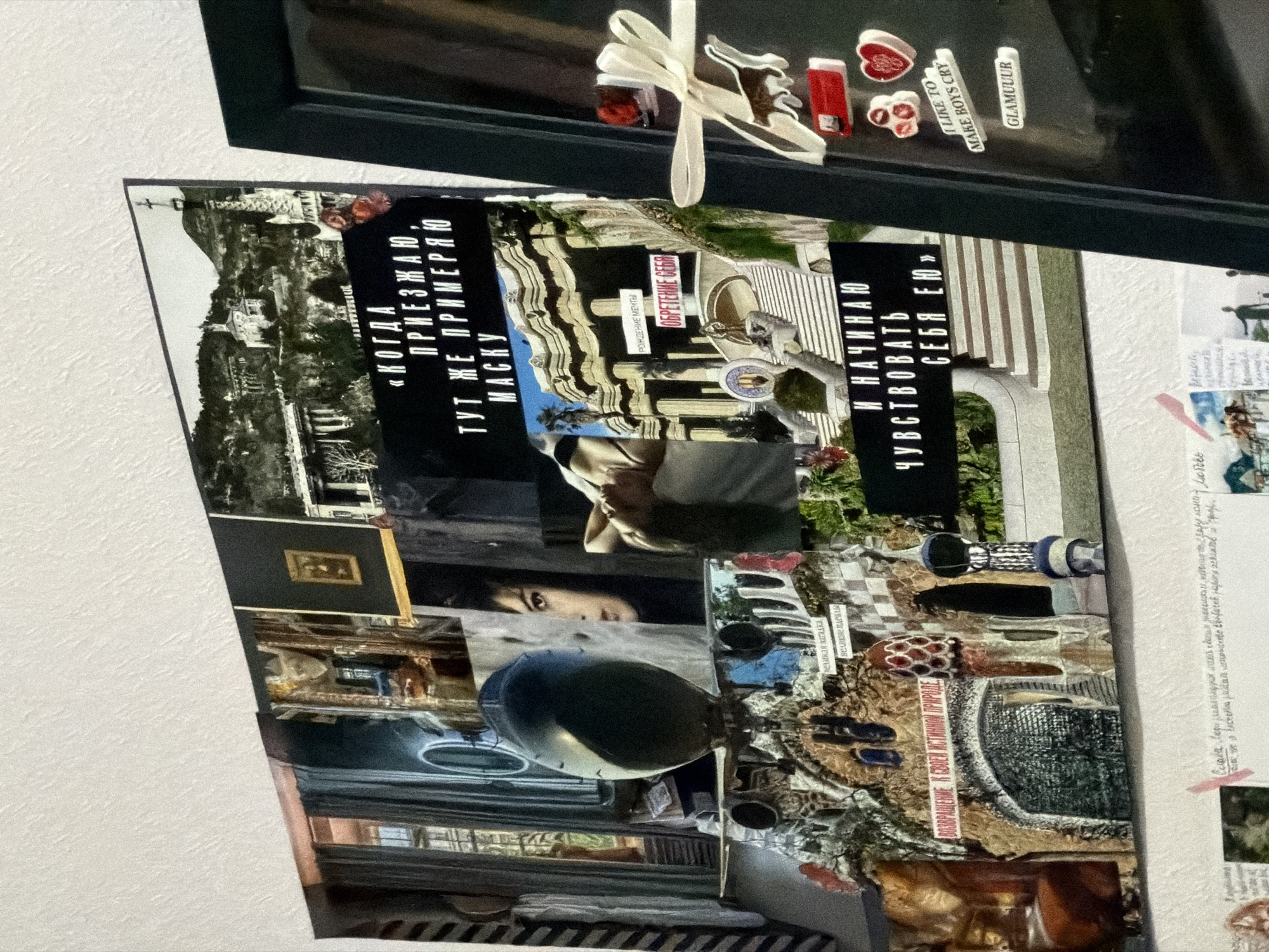 The image depicts a collage of images and text affixed to a white wall, with a dark framed object to the right. The collage features various photographs, including architectural structures, a close-up of a woman's face, a person lying down, and an image that appears to be a model airplane. Text in Cyrillic is overlaid on some of the images. The framed object on the right has several magnets attached to it, including a heart shape, a cat silhouette, and text that reads "I LIKE TO MAKE BOYS CRY" and "GLAMUUUR". The overall scene suggests a personal display of inspiration or memory. There is no identifiable indication of the city of Moscow, Russia, or any specific location within it.