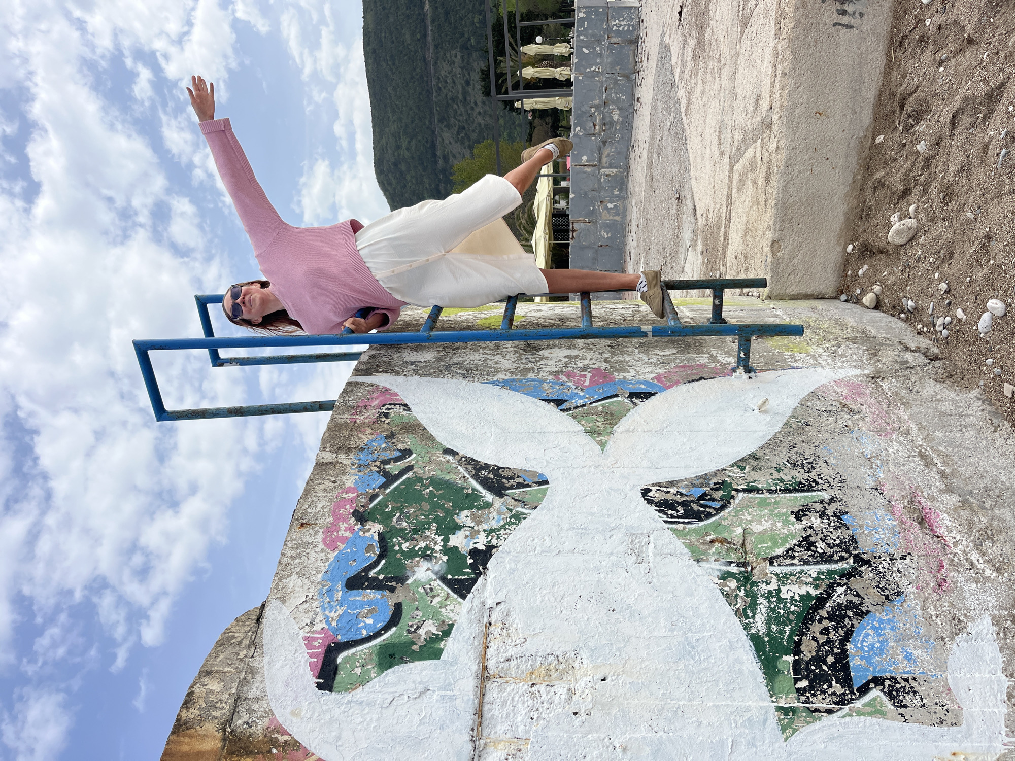 The image depicts a woman lying on a blue railing atop a concrete structure in what appears to be an outdoor setting. The woman is wearing a pink top, white shorts, and sunglasses. She is reclining with one arm raised towards the blue sky, which is filled with white clouds. Behind her is a mountain and some trees and a building in the distance.
The concrete structure beneath her is adorned with colorful graffiti. The environment around the concrete structure includes sand or dirt along the side of the structure. The photo may have been taken in or near Budva, Montenegro.