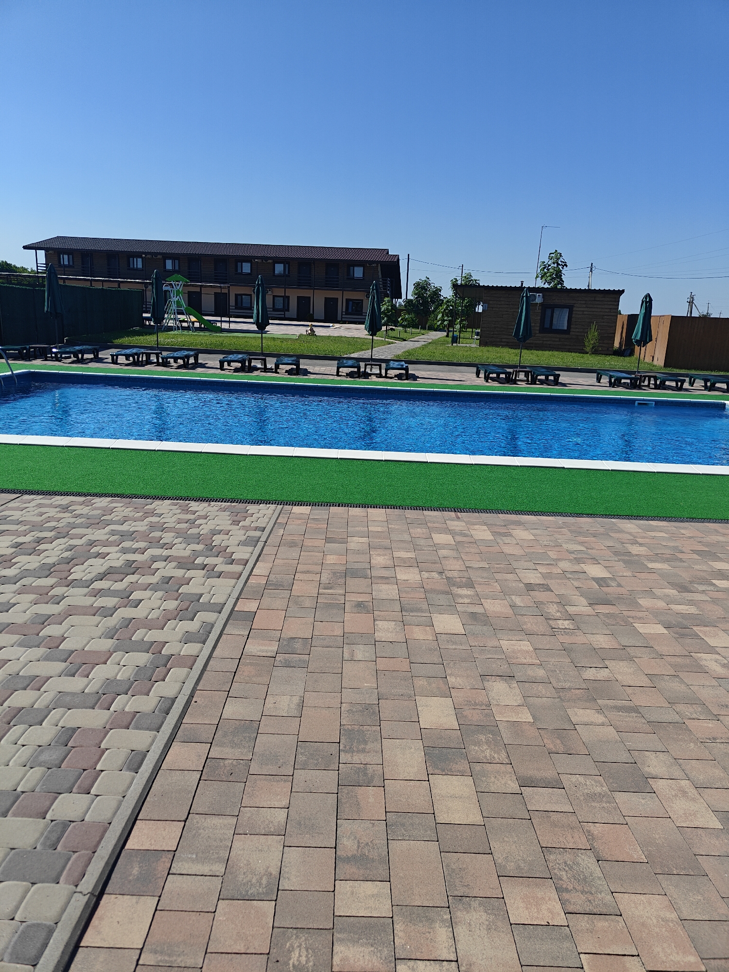 In Shkurupiivka, Ukraine, on a clear, sunny day, an outdoor swimming pool area is depicted. The pool itself is rectangular and filled with vibrant blue water, bordered by a white edge and a strip of artificial green turf. Beyond the turf, a paved walkway constructed from multi-colored interlocking pavers extends into the foreground.
To the left of the pool, several lounge chairs with dark blue cushions are arranged, some partially covered by green umbrellas. Further back, behind the pool and lounge chairs, a long, multi-story building with a dark brown roof and exterior siding is visible. This building has numerous windows and balconies. Adjacent to the building, a children's playground with a slide is present.
To the right of the main building, a smaller, wooden structure with a dark roof and a window can be seen, also shaded by green umbrellas. A wooden fence encloses part of the property in the background. The sky is a uniform, clear blue, with no clouds visible. The scene appears to be a resort or a recreational facility.