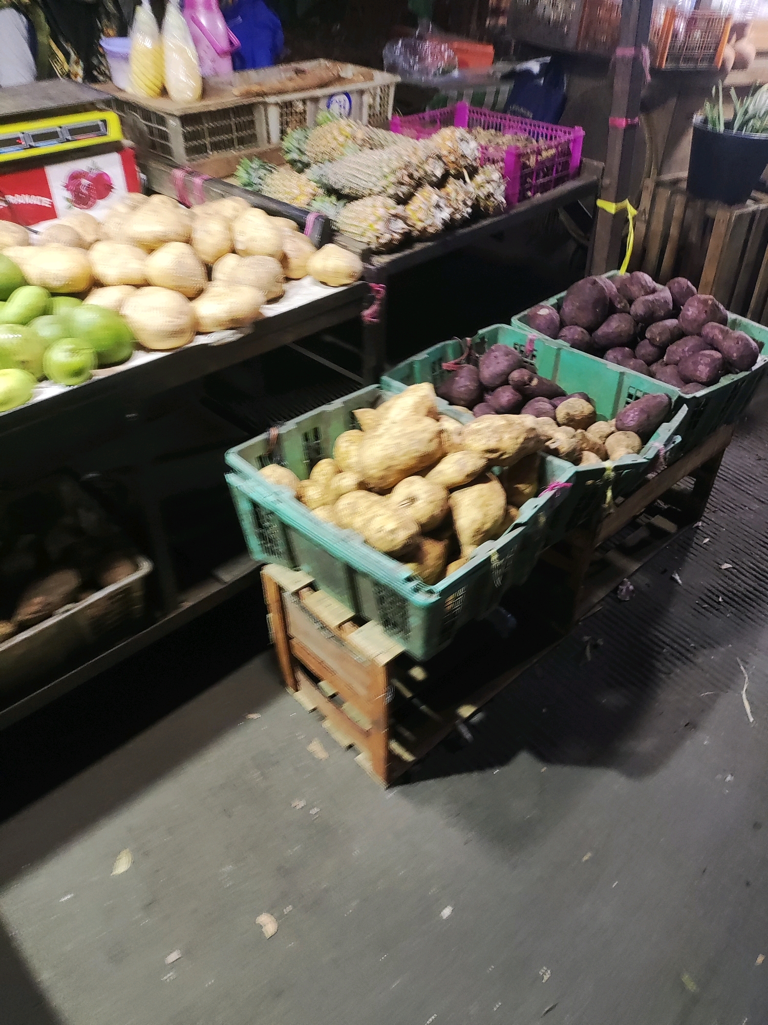 The image shows a market stall in East Jakarta, Indonesia, with a focus on various root vegetables and some fruits. In the foreground, two green plastic crates are filled with produce. The crate on the left contains light-colored, oblong root vegetables, possibly yams or sweet potatoes. The crate on the right is filled with darker, purple root vegetables, also likely a variety of sweet potato or yam. Behind these crates, a display of green apples can be seen on a black surface. Further back, a pink plastic crate holds pineapples, and a wooden crate with a black plastic insert contains more of the dark purple root vegetables. In the background, there are various other market items and structures, including what appears to be a weighing scale and some bagged liquids. The overall scene depicts a bustling market environment.