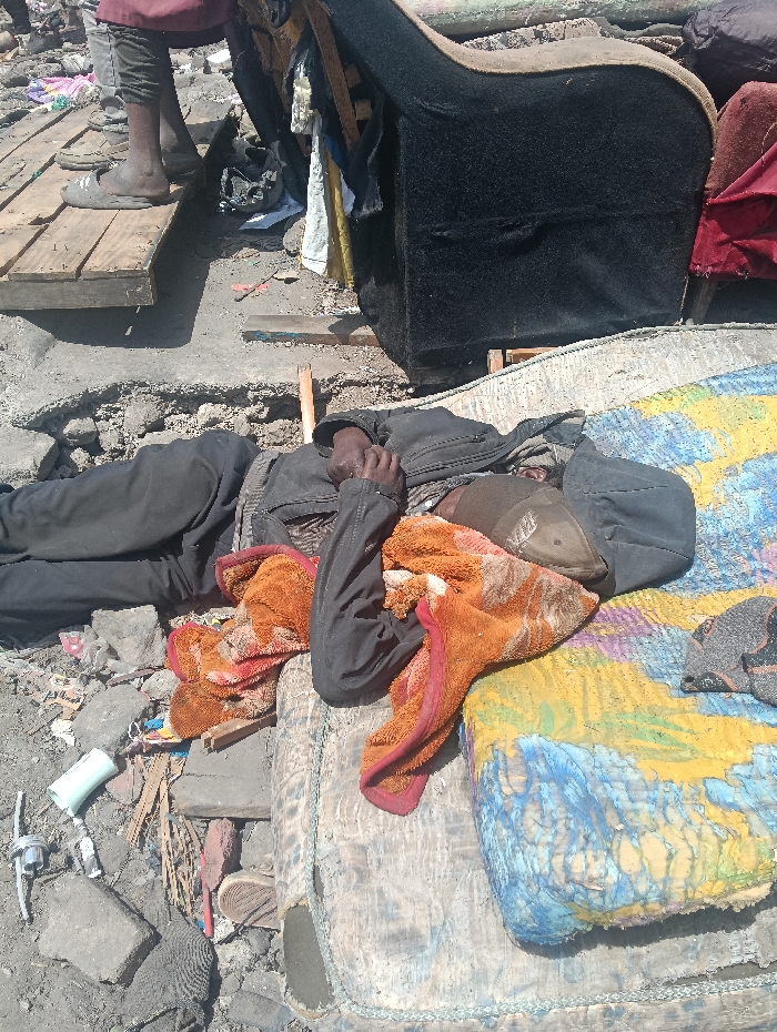 A person lies on a tattered mattress, covered with a worn, orange blanket and wearing a black jacket and hat. The scene is surrounded by debris and trash, including broken pieces of concrete, discarded items, and remnants of furniture. A pallet-like platform is nearby. The overall environment suggests poverty and destitution.