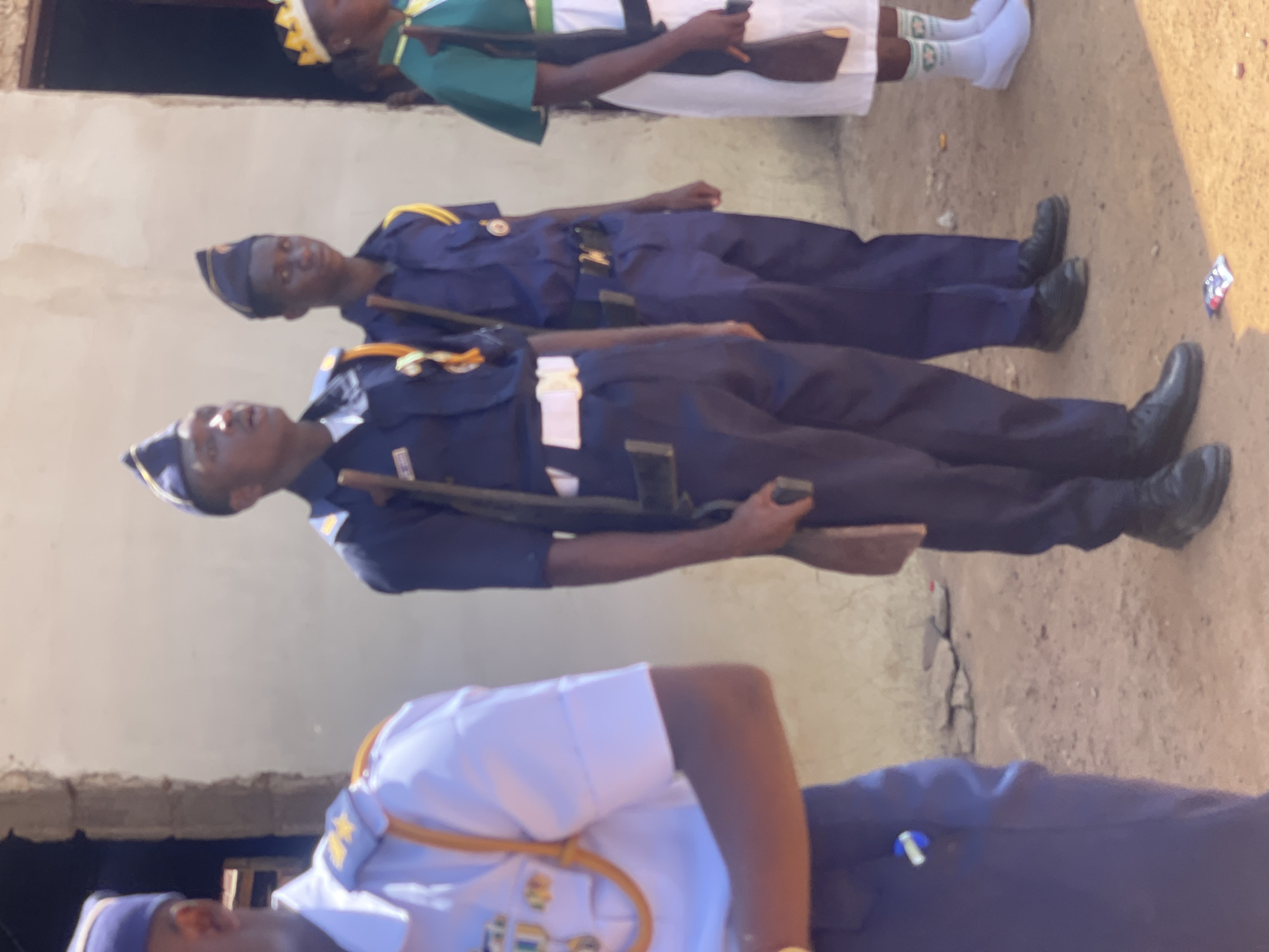 The image, oriented vertically, captures a group of individuals, predominantly young people, standing outdoors in what appears to be a formal or ceremonial setting in Andaha, Nigeria.
In the foreground, partially visible, are two individuals. One wears a white uniform shirt with gold trim on the collar and epaulets, a dark tie, and a dark cap with a gold emblem. The other, mostly out of frame, wears a dark blue uniform.
Behind them stand two young men in full dark blue uniforms, complete with gold-trimmed epaulets and dark blue caps with emblems. They both hold long, slender wooden staffs or mock rifles, positioned vertically at their sides. They are wearing black shoes. On the light brown, dirt ground near their feet, a crumpled piece of litter, possibly a wrapper, is visible.
Further back and slightly elevated, a young woman is seen. She is dressed in a white gown or dress, adorned with a green sash, and wears a distinctive yellow and white patterned head covering, resembling a crown. She has white socks with green patterns and white shoes. She appears to be holding a dark brown object.
The background consists of a plain, light-colored wall, possibly beige or light brown, suggesting an architectural structure. Above the wall on the left, a darker, shadowed area might indicate a window or opening. The lighting suggests it is daytime, with bright sunlight illuminating the scene, indicative of clear weather. The individuals' expressions appear focused or neutral, consistent with participation in a formal event, possibly a parade or youth group gathering. No discernible text is visible in the image.