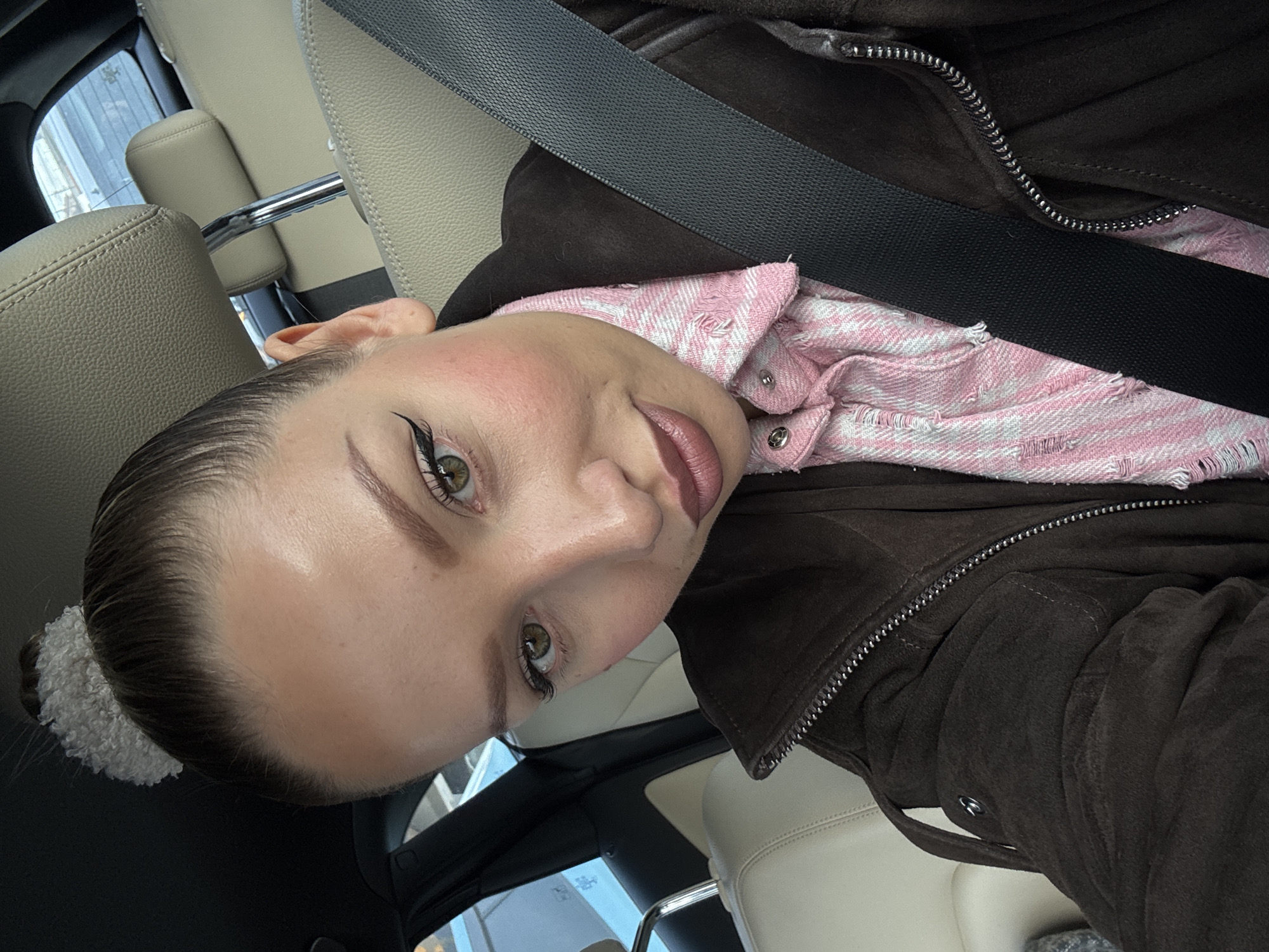 A close-up selfie shows a person with light skin, green eyes, and dark winged eyeliner, looking directly at the camera. The person's hair is pulled back tightly into a bun, secured with a light, fluffy, possibly white or cream-colored scrunchie. Prominent blush is visible on the cheeks, and the lips are colored with a dark pink or mauve lipstick.
The person is wearing a dark brown, possibly suede, jacket with a visible silver zipper running down the front. Beneath the jacket, a pink and white striped shirt with frayed or distressed edges and silver snap buttons is visible. A black seatbelt diagonally crosses the person's chest.
The setting appears to be the interior of a car, indicated by light-colored (beige or cream) leather seats visible in the background and partially in the foreground. A window reflection in the background shows an indistinct outdoor view, suggesting it is daytime. The image itself is rotated 90 degrees counter-clockwise, making the person's head appear tilted. There is no visible text.