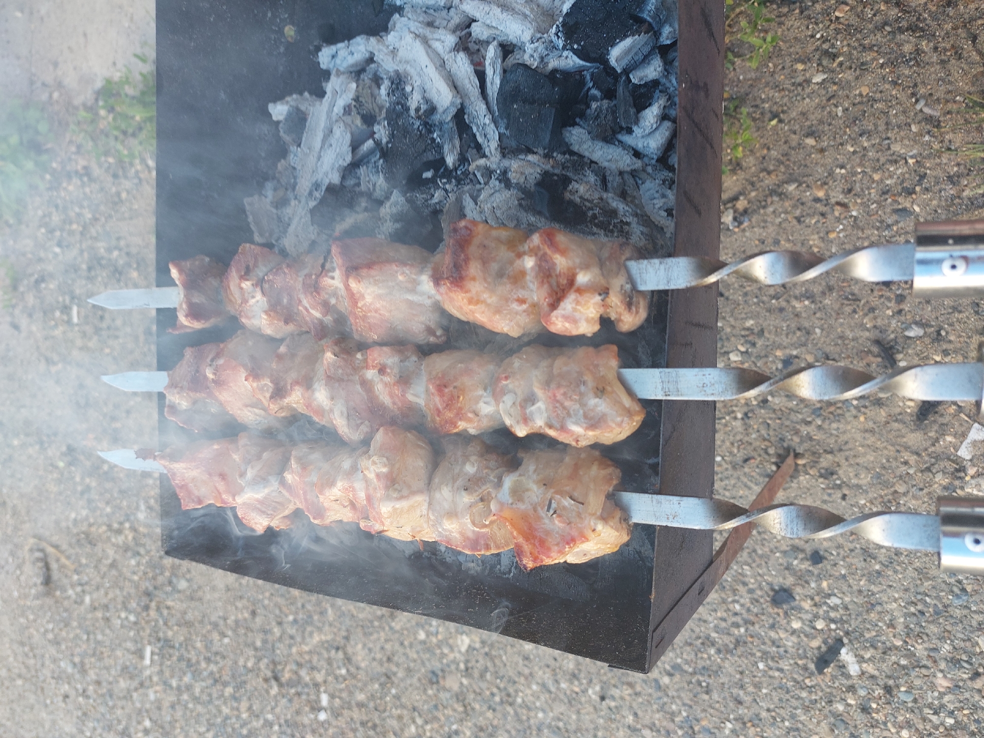 A close-up, eye-level, outdoor shot captures three skewers of raw meat cooking over glowing coals in a metal grill. The meat, cut into uniform cubes, is arranged on metal skewers, with each skewer holding approximately six pieces. Smoke rises from the grill, obscuring the lower half of the meat and creating a hazy atmosphere. The grill itself is dark metal, positioned on a gravelly surface. The background consists of more gravel and sparse green vegetation, suggesting a rural or park-like setting. The scene is devoid of any human presence. The image does not provide any information about the location in the city of Chaikovsky, Russia.