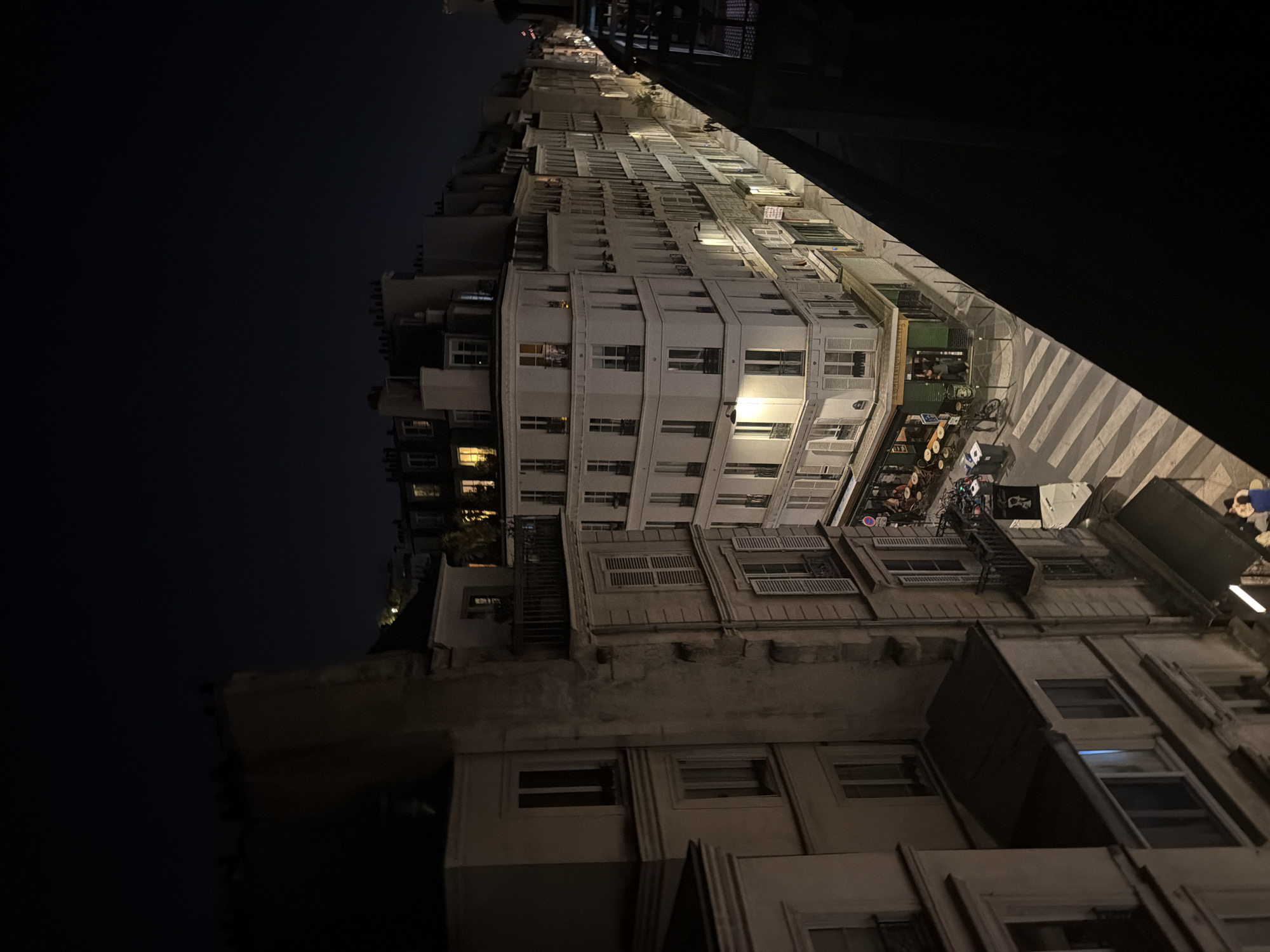 At night, the image showcases an array of multi-story buildings in Paris, France. The architectural style includes white facades with regularly spaced windows. Some windows are adorned with shutters, and fire escapes are visible on some buildings. The street below is illuminated, revealing a zebra crossing and people gathered near a shop or café. The environment is a mix of light and shadow, with an urban atmosphere. The buildings are densely packed, characteristic of Parisian urban design.