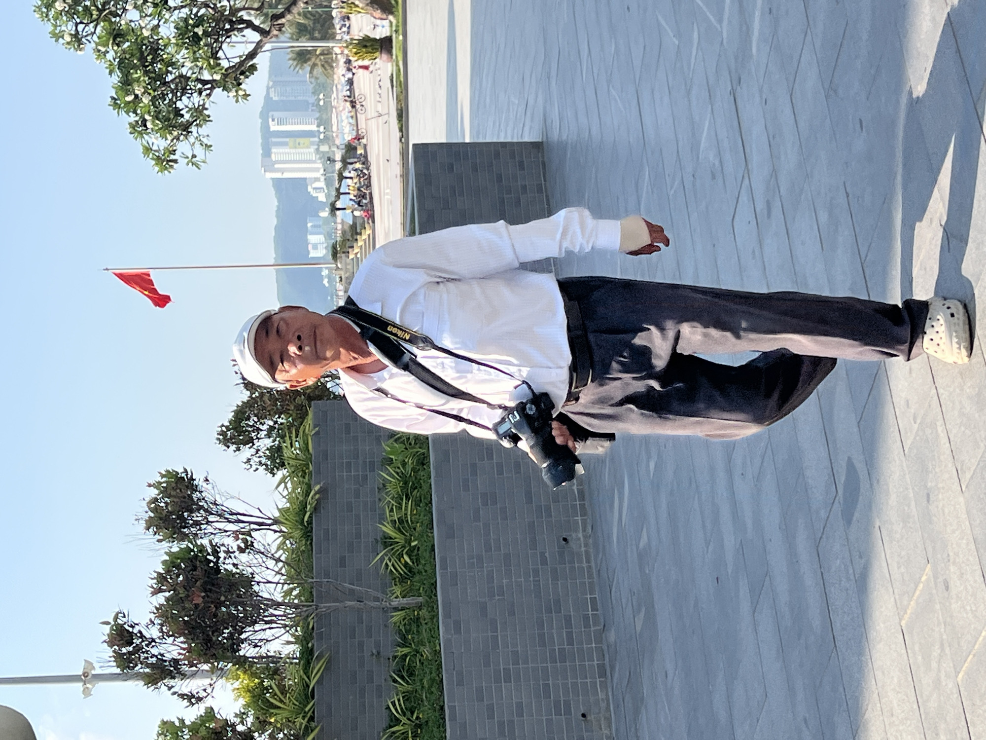The image shows an Asian man standing on a paved surface. He is wearing a white long-sleeved shirt, dark trousers, and white Crocs. A black camera with a Nikon strap hangs around his neck. He has a white cap on his head and a bandage on his right hand. The background includes trees, plants, a gray brick wall, a red flag, and buildings in the distance. Based on the context clues, the location is likely Nha Trang, Vietnam.
