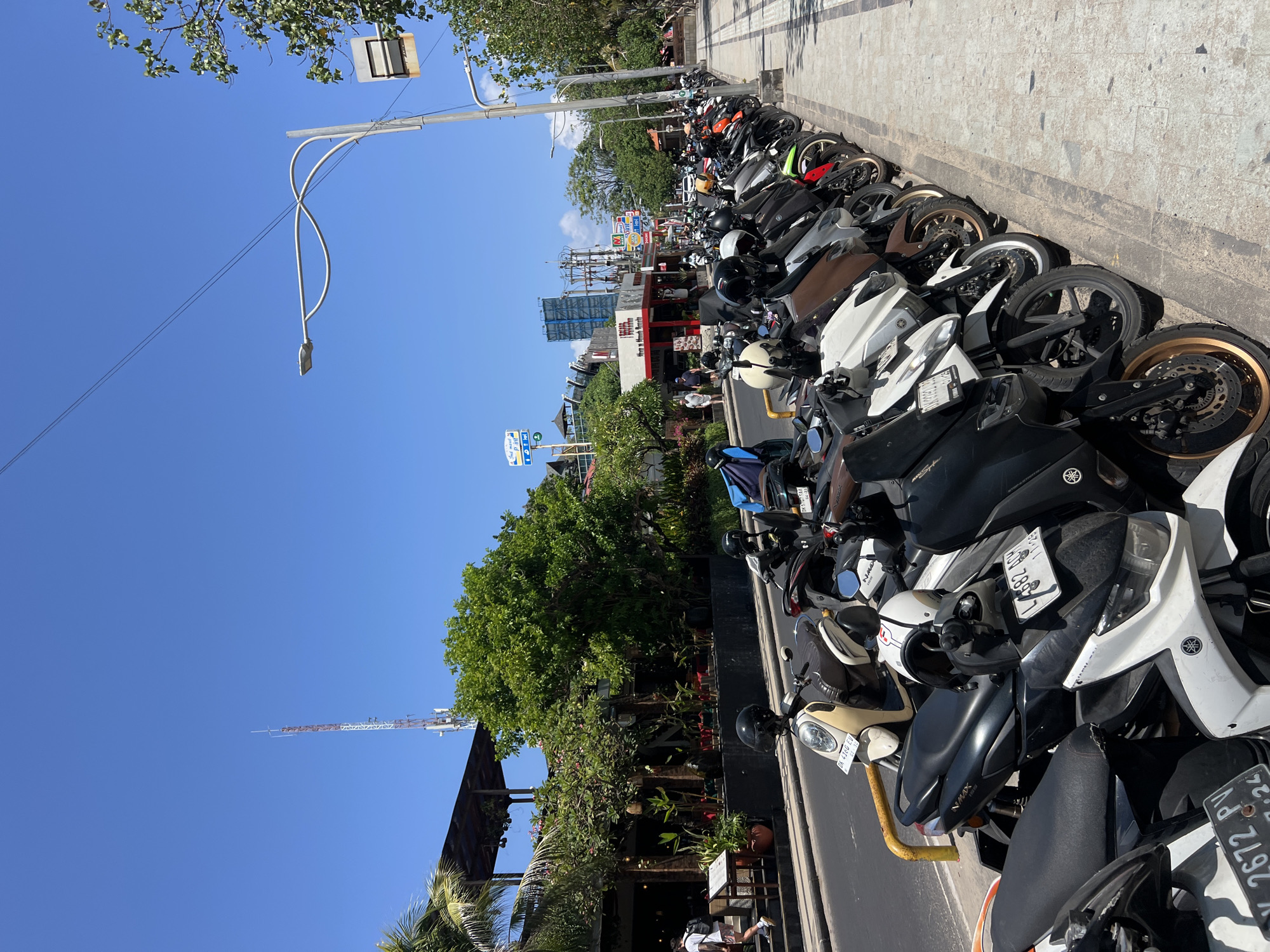 The image depicts a street scene in Kuta, Indonesia, on a clear, sunny day with a bright blue sky. A row of parked scooters and motorcycles lines the right side of the image, parallel to a paved sidewalk. The vehicles are of various colors and models, many with helmets placed on their seats or handlebars. Beyond the sidewalk, a street is visible with a few cars and more motorcycles in motion. On the left side of the street, there are buildings with outdoor seating areas, lush green trees, and some palm trees, suggesting a commercial or dining district. In the background, a telecommunications tower and several other buildings are visible. A street light with a distinctive curved design is prominent in the upper right portion of the frame. The overall scene suggests a busy urban environment, characteristic of a popular tourist destination.