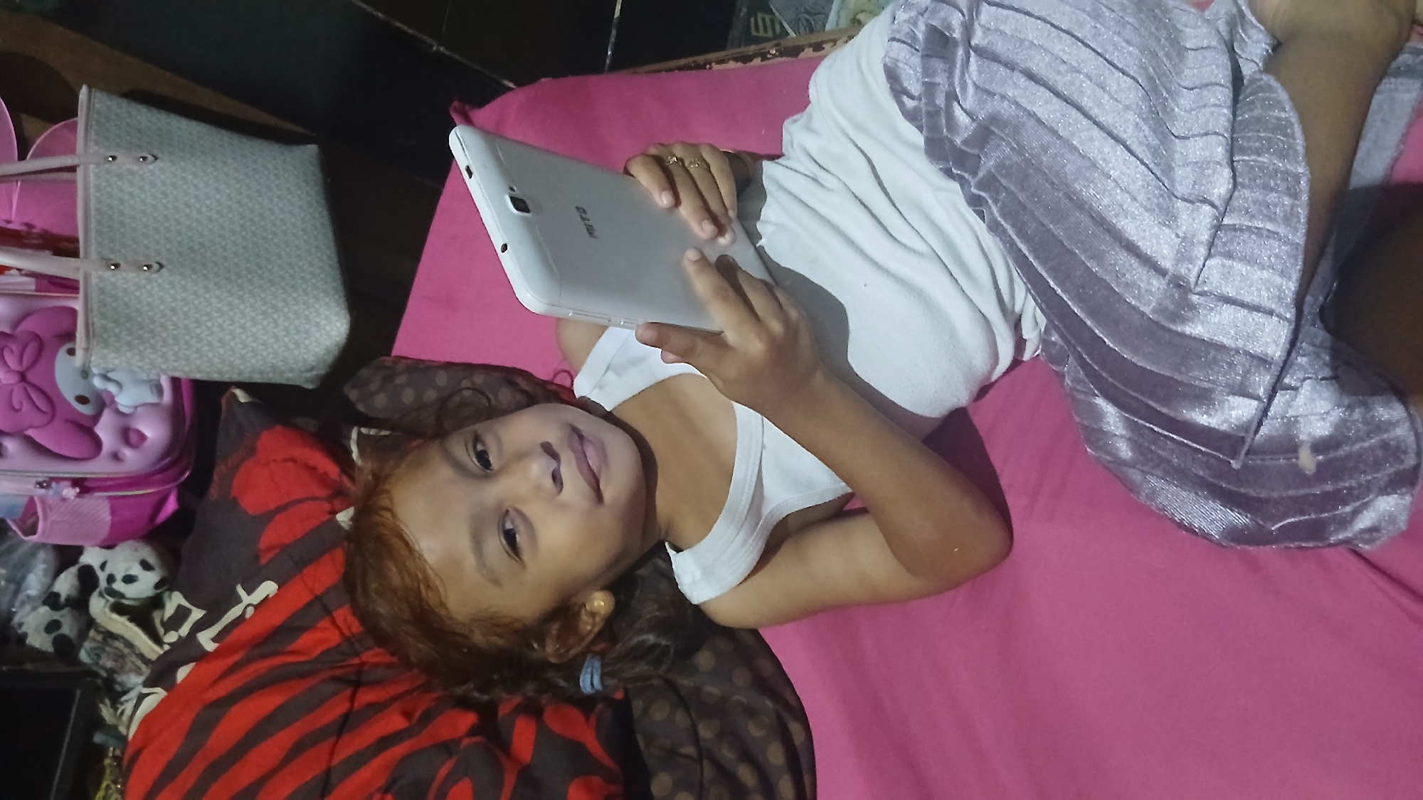 A young girl is lying on a pink surface, possibly a bed, holding a white tablet computer. She is wearing a white tank top and light purple, pleated pants. Her hair is reddish-brown and tied back with blue hair ties. In the background, a pink backpack with a cartoon character design and a light-colored tote bag are visible. There are also pillows with red and black striped and brown polka dot patterns. The setting appears to be an indoor environment in Keudejungkagajah, Indonesia.
