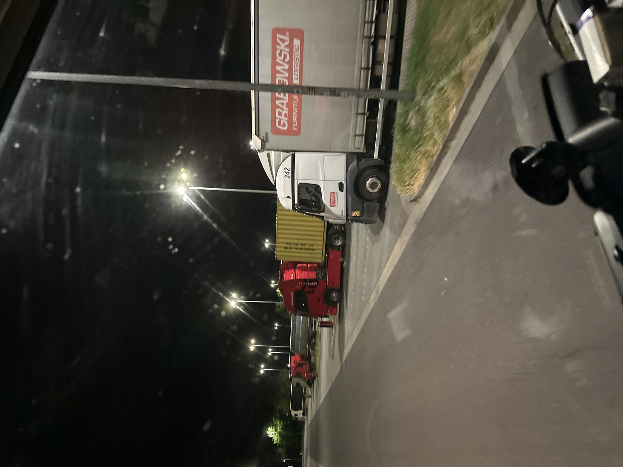 A nighttime scene captures several parked semi-trucks on a paved lot, likely a rest stop or truck yard, located in Luckau, Germany. The foreground is dominated by the interior of a vehicle, with a black dashboard and steering wheel visible. In the midground, multiple semi-trucks are lined up. The closest truck is white with a large grey trailer bearing the red logo "GRABOWSKI FURNITURE LOGISTICS." To its left are two red semi-trucks, one with a yellow container. Further down the line, another white truck is visible. The background consists of streetlights illuminating the area, trees, and a dark sky. The ground is asphalt, with some patches of grass along the edge.