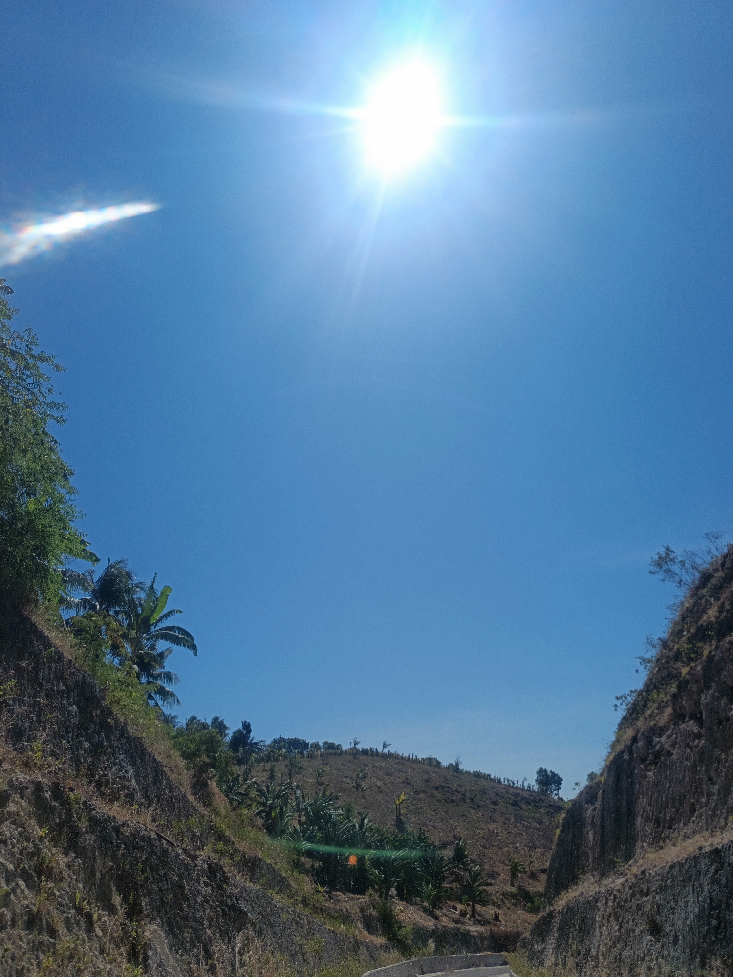 This image depicts a bright, sunny day in a rural area, possibly within the city of Gorontalo, Indonesia. The foreground features a road cutting through a landscape dominated by steep, rocky hillsides. The right side of the image shows a sharply cut cliff face with a textured, rocky surface, revealing layers of sediment. Sparse, dry vegetation clings to the slopes.
On the left side, another rocky hillside rises, also sparsely vegetated with dry grasses and some greenery. Lush banana trees and other tropical foliage are visible on the slopes and in the midground, indicating a warm climate. The sky is a clear, vibrant blue, with the sun intensely bright and high in the sky, creating visible lens flares and starburst effects. A faint, elongated streak of light, possibly a contrail or atmospheric phenomenon, is visible in the upper left portion of the sky. The overall impression is one of a dry, sun-drenched tropical environment, likely a rural or semi-rural outskirts area. There are no people or animals visible in the frame.