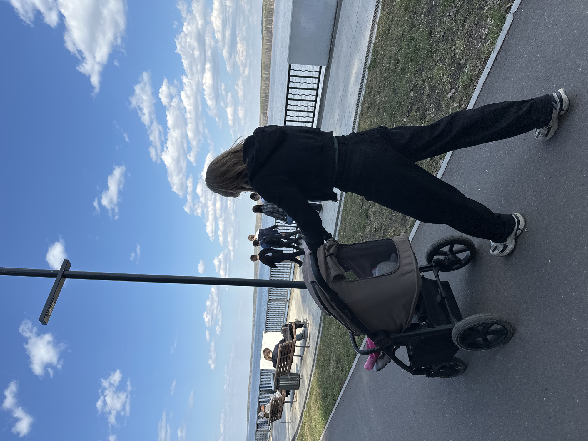 A woman in black clothing and athletic shoes pushes a baby stroller along a paved walkway. The image is taken from an upward, off-center angle, making the scene appear tilted. The walkway runs alongside a body of water, possibly a lake or wide river, with a metal railing separating the walkway from the water's edge. Benches line the walkway, and several people are seated. Other people walk along the path in the background. The sky is bright blue with scattered white clouds. A street lamp with a small sign is visible on the left. The scene is identified as being in the city of Chaikovsky, Russia.