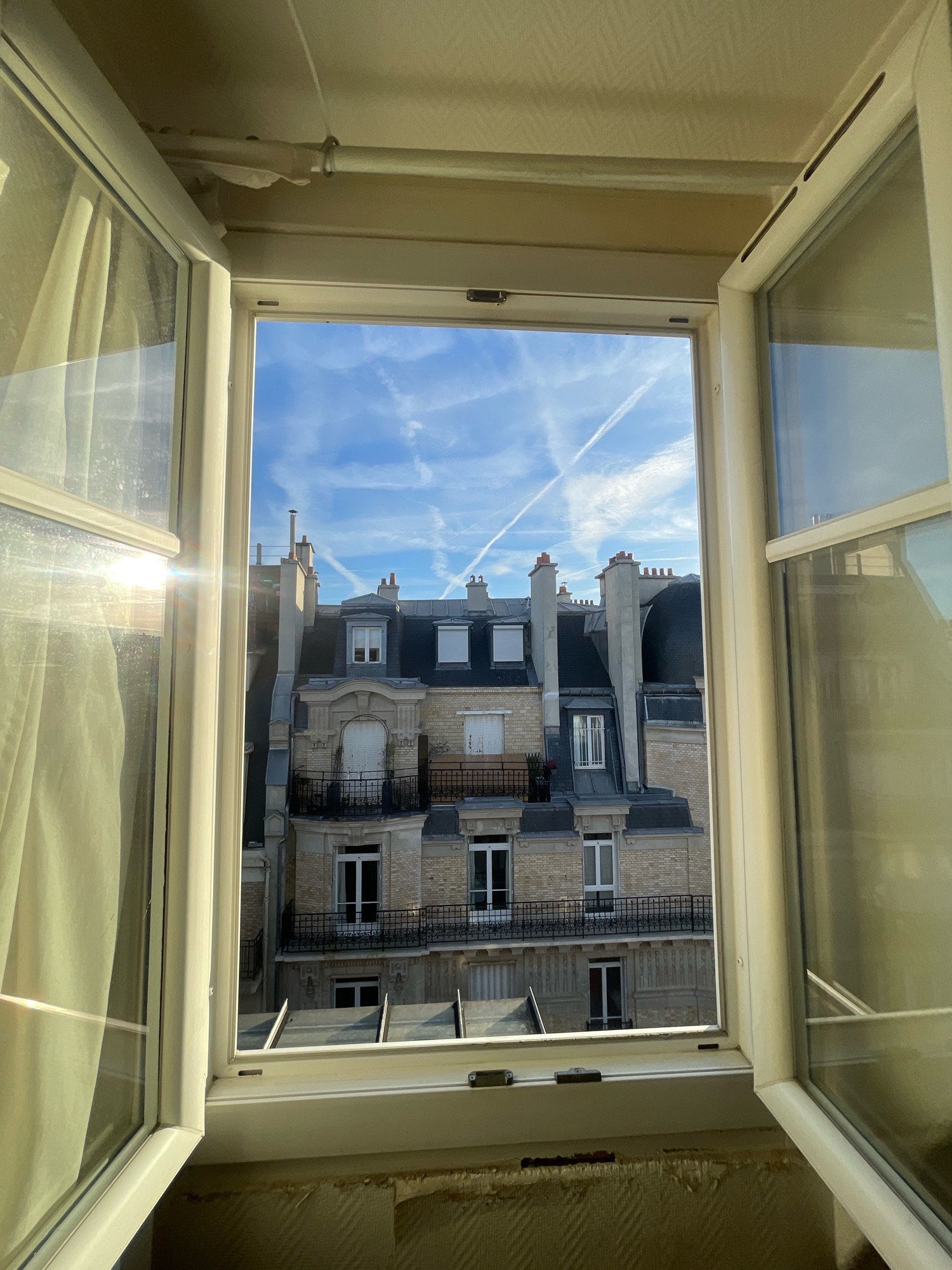 This image captures a view through an open window in Paris, France, overlooking a Parisian cityscape. The window itself is white and slightly ajar, with one pane reflecting bright sunlight and a cream-colored curtain to the left. Through the window, a traditional Parisian building with ornate architectural details, multiple windows, and balconies with black wrought-iron railings is visible. The building's facade is a combination of light-colored brick and stone. Above the building, a bright blue sky is dotted with wispy white clouds, some forming contrails from aircraft. The scene is illuminated by natural daylight, suggesting it is daytime. The interior framing of the window and the wall below are also visible, showing a textured wallpaper.