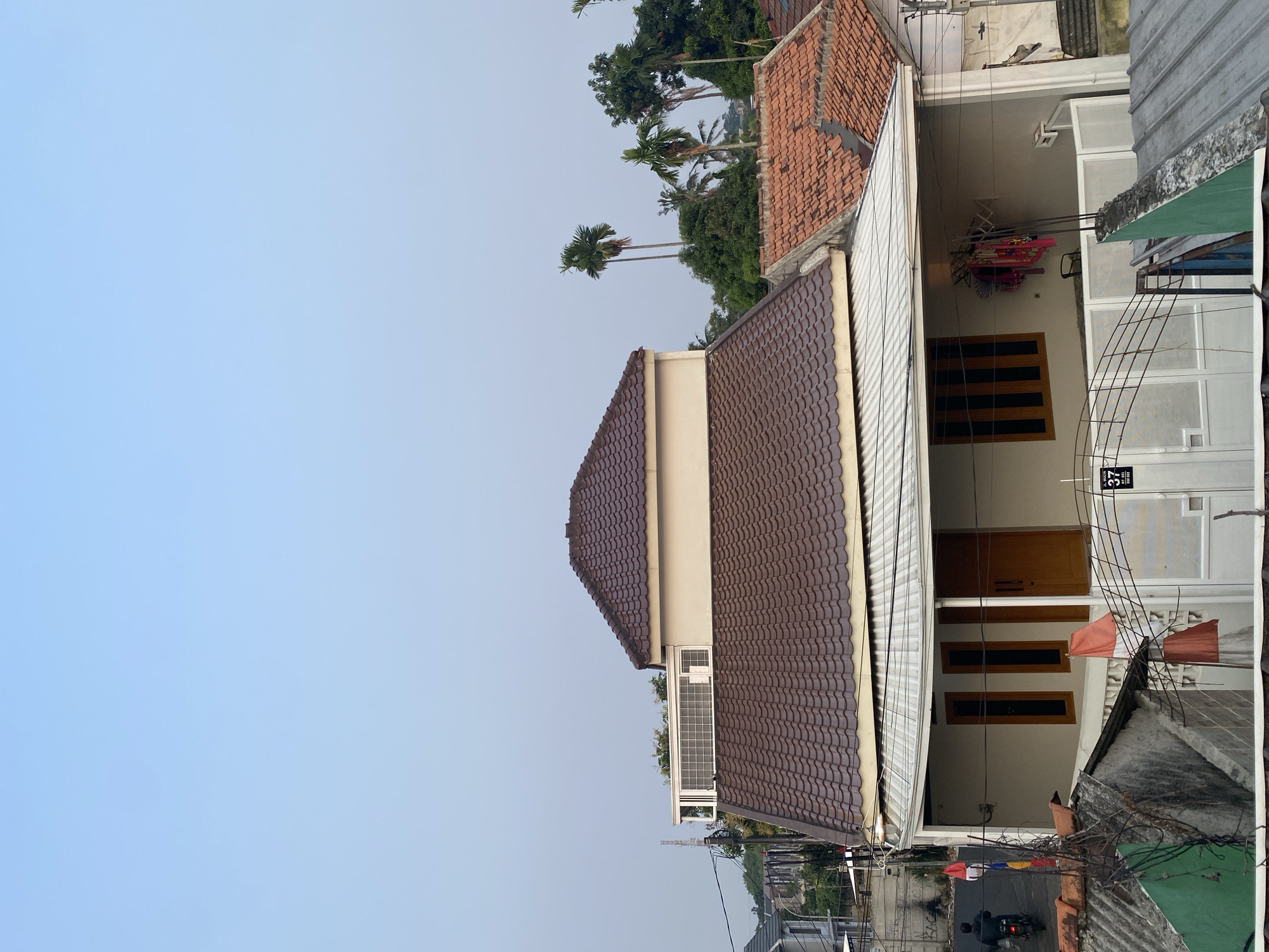 The image is an exterior view of a residential house in Tangerang, Indonesia. The house has a light-colored facade with a brown tiled roof. A second story with a similar brown tiled roof is visible above the main roofline. The house has several windows with wooden frames and a wooden front door. A covered porch area extends from the front of the house, with a clothesline holding laundry. A white metal gate with decorative elements is in front of the porch. A sign with the address "37 JL. WIJAYA RT. 183 RW. 002" is attached to the gate. The surrounding environment includes other houses with tiled roofs, trees, and utility poles. In the background, a motorcycle rider is seen on a street. The sky is clear and light blue, suggesting it is daytime.