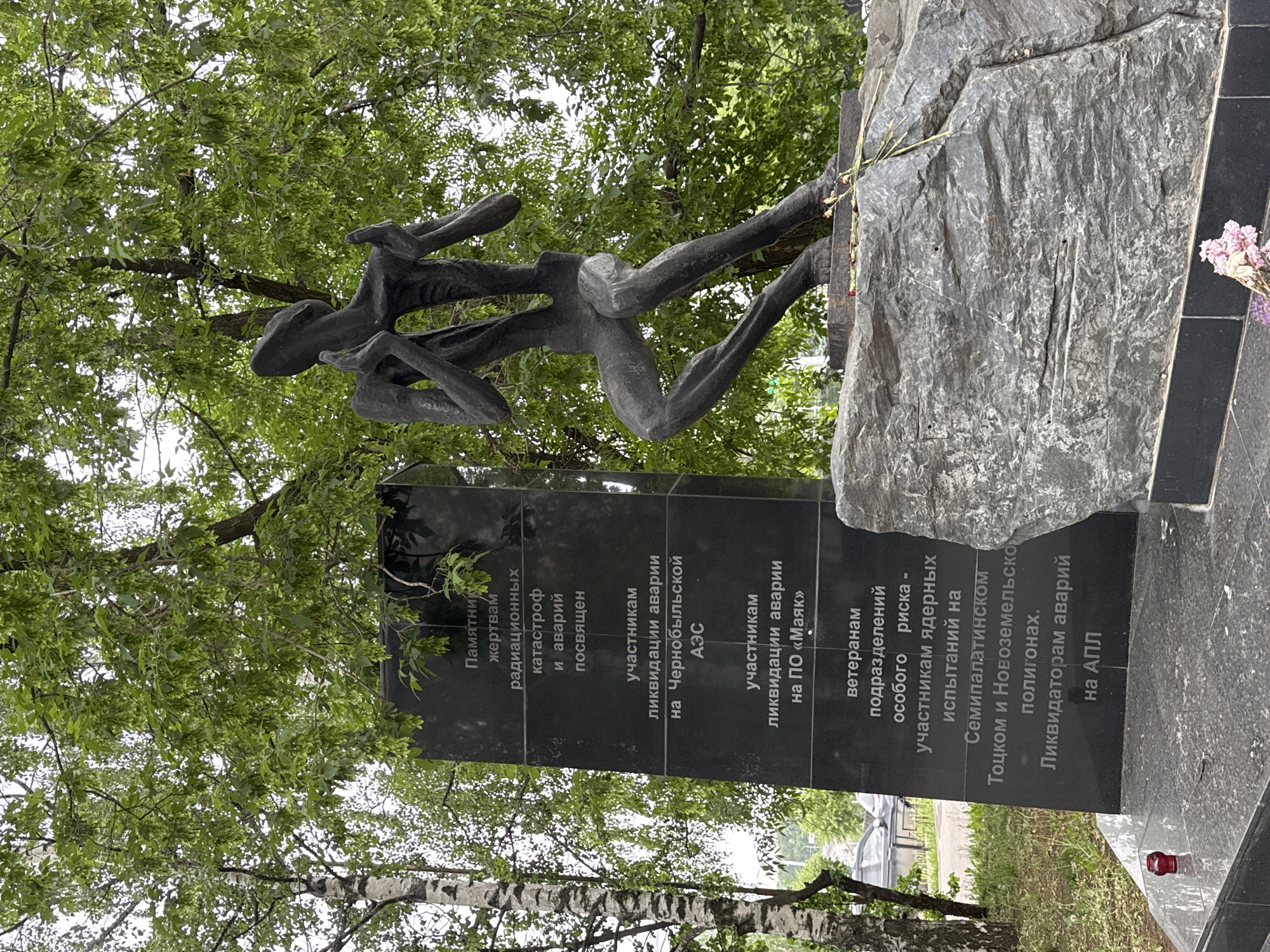 The image depicts a monument in Izhevsk, Russia, dedicated to victims of radiation-related catastrophes and accidents. The monument consists of a black granite stele with inscriptions in Russian and a bronze sculpture of abstract human figures. The inscriptions mention the Chernobyl accident, the "Mayak" production association, veterans of special risk units who participated in nuclear tests in Semipalatinsk, Totsk, and Novaya Zemlya test sites, and liquidators of accidents on nuclear submarines. The monument is surrounded by greenery, suggesting an outdoor park setting. Flowers are placed near the monument, possibly in remembrance.