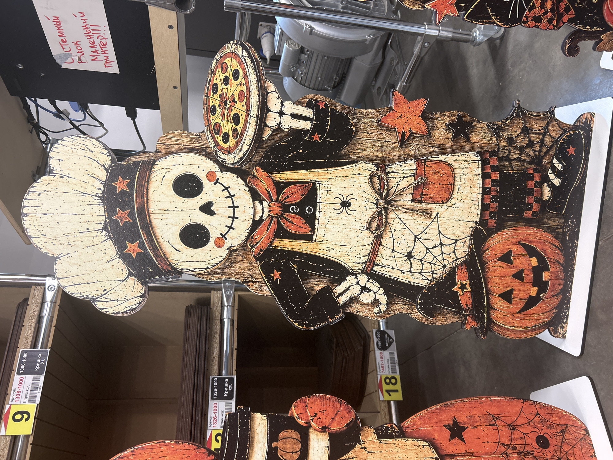 The image shows a Halloween-themed cutout of a skeleton chef holding a pizza. The cutout is made of wood and has a rustic, vintage appearance. The skeleton is wearing a chef's hat, apron, and checkered pants. A jack-o'-lantern and a black cat are also visible in the background. The scene appears to be set up in a retail store, possibly a Halloween decoration aisle. There are shelves with products in the background, and price tags are visible. A sign written in red marker on a black surface reads "Системный блок маленький принтер!!!" which translates to "System unit small printer!!!". There is no indication of the specific location in Ternopil, Ukraine.