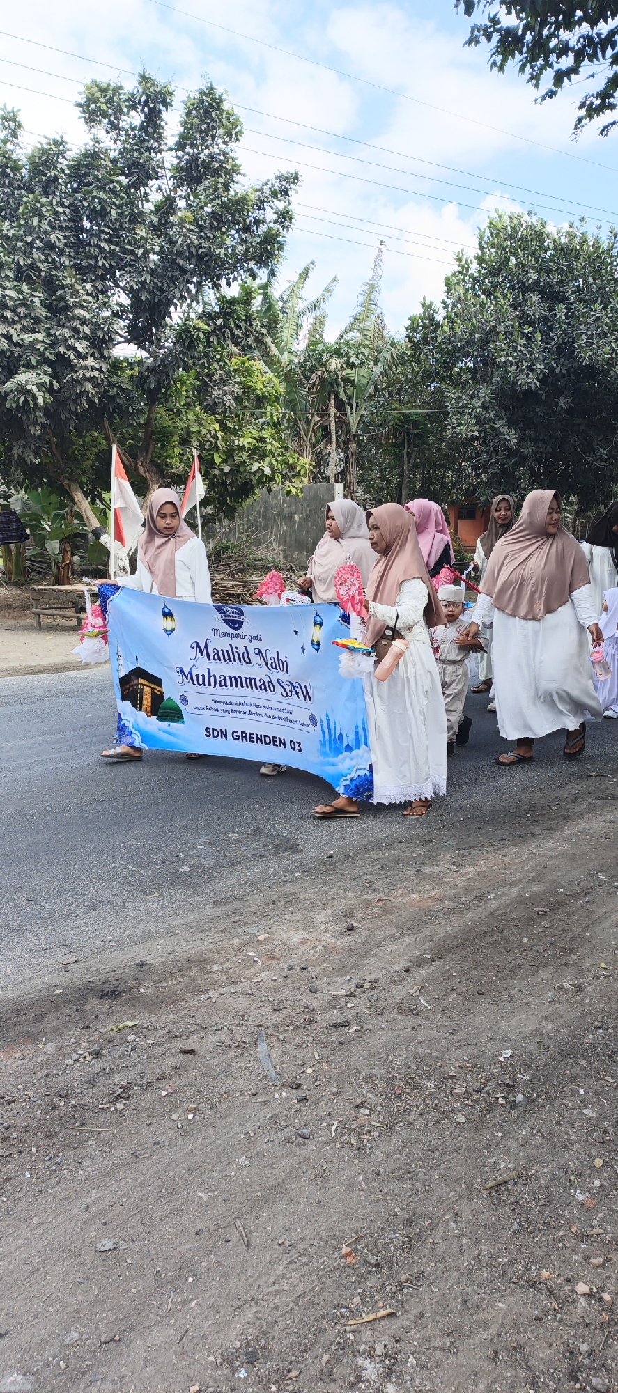 In Grenden, Indonesia, on a paved road under a cloudy sky, a group of people, primarily women and children, are participating in a procession. They are walking alongside a large banner that reads "Memperingati Maulid Nabi Muhammad SAW" and "SDN GREDEN 03". The banner features images of the Kaaba and mosque domes. The participants are dressed in Islamic attire, with many wearing hijabs. Some are carrying decorative lanterns and a water bottle. The environment appears to be a rural or semi-urban area, with trees and a low wall visible in the background. The ground is a mixture of asphalt and gravel.