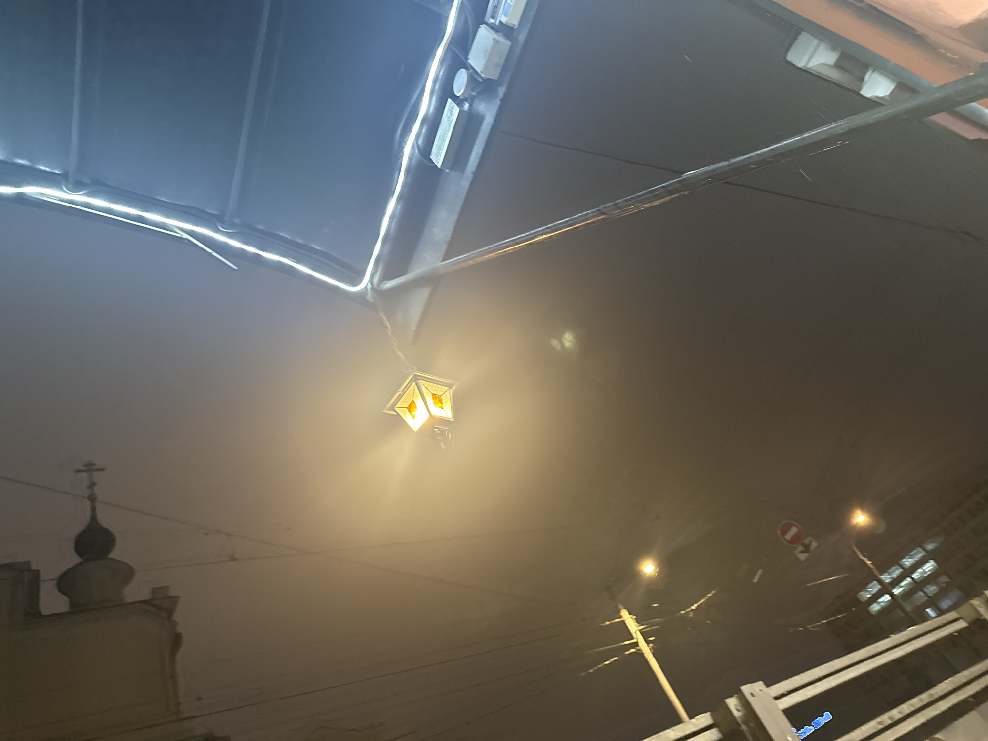 The image captures a night scene in Moscow, Russia, characterized by heavy fog or smog that obscures distant views. The foreground is dominated by an overhanging structure, possibly a canopy or roof extension, illuminated by linear LED strip lighting. A decorative street lamp with a cage-like design and warm yellow light is suspended from the structure. In the background, a church steeple with a cross is partially visible through the fog. Streetlights and power lines are also discernible, contributing to the urban setting. A fenced area is present at the bottom right, and a road sign can be seen, indicating a pedestrian crossing or restricted zone. The overall atmosphere is dim and misty.