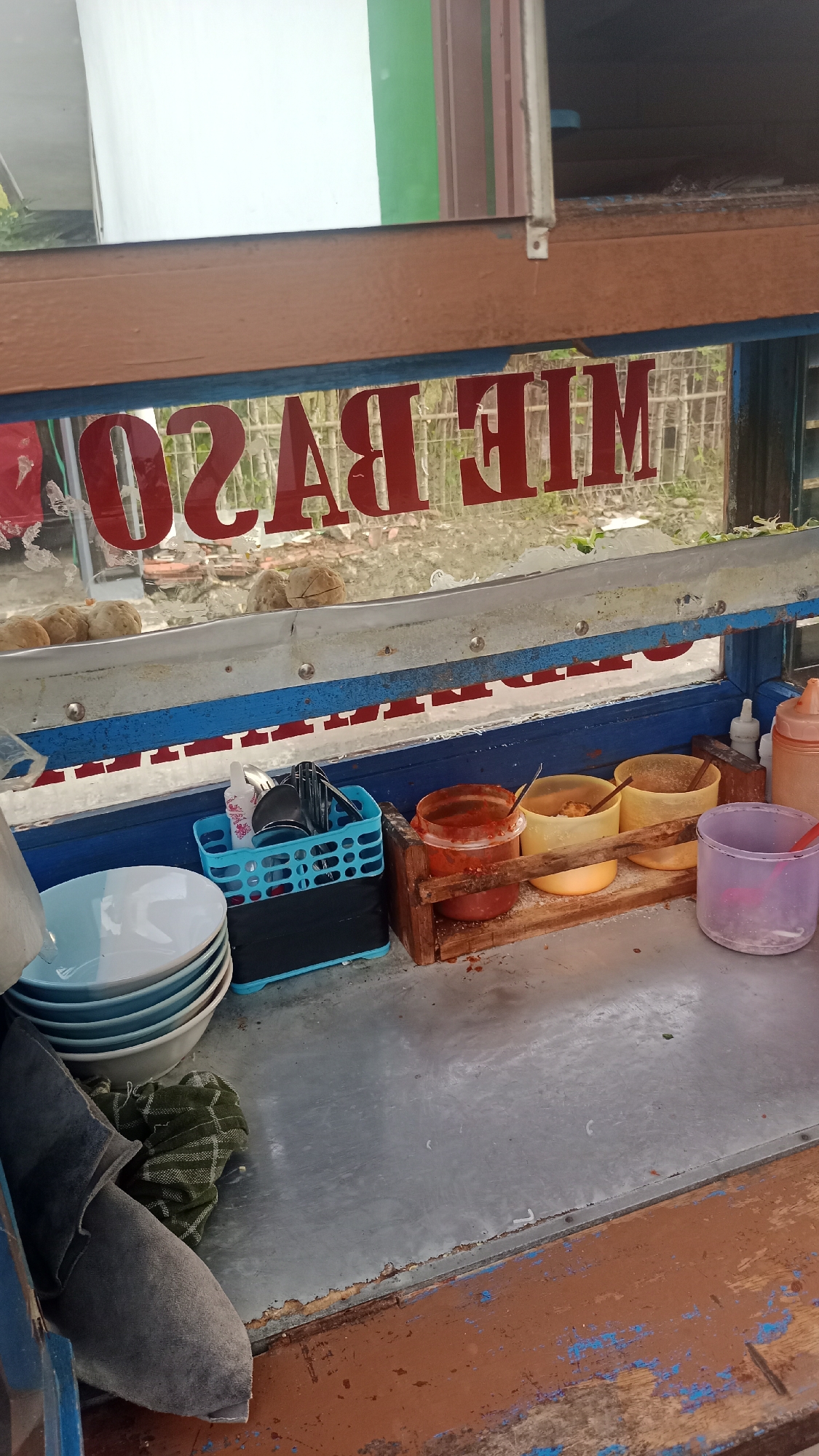 The image captures a close-up view of a food cart, likely selling "mie baso" (noodle soup with meatballs), as indicated by the red text on the window. The cart appears to be located outdoors, possibly in the city of Pakemitan, Indonesia, given the context.
The foreground is dominated by a metal countertop, showing signs of wear and tear with peeling paint. On the left, a stack of light blue and white bowls is visible, along with a folded green and white checkered cloth and what appears to be a dark gray fabric or apron.
To the right of the bowls, a blue plastic basket holds various utensils, including spoons and forks, along with a smaller white cup. Adjacent to the basket is a wooden holder containing three small containers. The leftmost container holds a red sauce or chili paste, the middle one appears to contain yellow seasoning or crisps, and the rightmost one is empty. A purple plastic cup with a red utensil is also present on the far right.
Behind the countertop, a window displays red lettering that reads "MIE BASO". Through the window, a blurry background shows what seems to be a yard with some greenery and possibly some structures in the distance. A few round, beige objects, likely meatballs, are visible lined up on a ledge above the window. A bottle with an orange lid, possibly a condiment dispenser, is positioned on the far right side of the cart.
