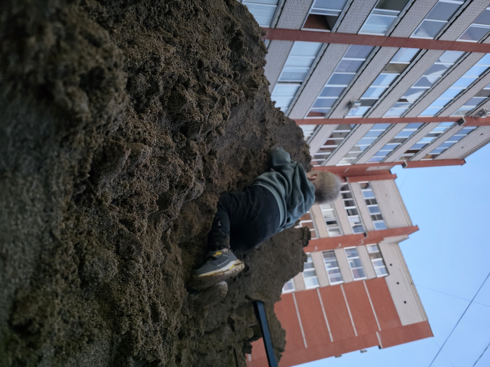 In Chaikovsky, Russia, a low-angle shot shows a young child, seen from behind, climbing a large mound of dark sand. The child is wearing a green hooded sweatshirt and dark pants, with one shoe visible on the sand. In the background, two modern apartment buildings rise against a clear blue sky. One building is predominantly gray with red accents and numerous windows, while the other is red with cream-colored sections and similar windows. The scene appears to be an outdoor public space, possibly a playground, given the sand mound and the presence of apartment buildings, common in urban Russian environments. The time of day is likely late afternoon or early evening due to the soft lighting.
