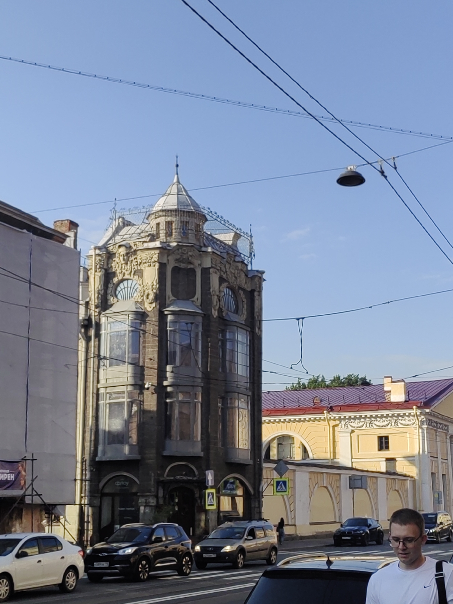 The image captures a street scene in Saint Petersburg, Russia, on a clear, sunny day, likely in the late afternoon or early evening given the long shadows. The foreground features a man wearing glasses and a white t-shirt, carrying a bag over his shoulder, and looking downwards with a neutral expression. He is standing near a black car, with other vehicles, including a white sedan and a dark SUV, visible on the street.
Dominating the mid-ground is an ornate, historic building with a unique architectural style, possibly Art Nouveau, characterized by its elaborate facade, bay windows, and a distinctive turret-like structure at the top. The building is dark in color with lighter decorative elements. Adjacent to it is a yellow building with a reddish-purple roof, featuring arched windows and classical architectural details.
The background shows a clear blue sky with overhead wires and a street lamp. Some greenery is visible above the yellow building, suggesting trees. The overall scene depicts an urban environment with a blend of historic and modern elements, including traffic and pedestrians.
There are a few instances of visible text: a sign on the left side of the image appears to have Russian lettering, with "ИМРЕН" partially visible. Another sign on the dark building has the word "Фрэнси" or similar in Cyrillic. A pedestrian crossing sign is also visible. The presence of Cyrillic text strongly supports the Saint Petersburg, Russia location context.
