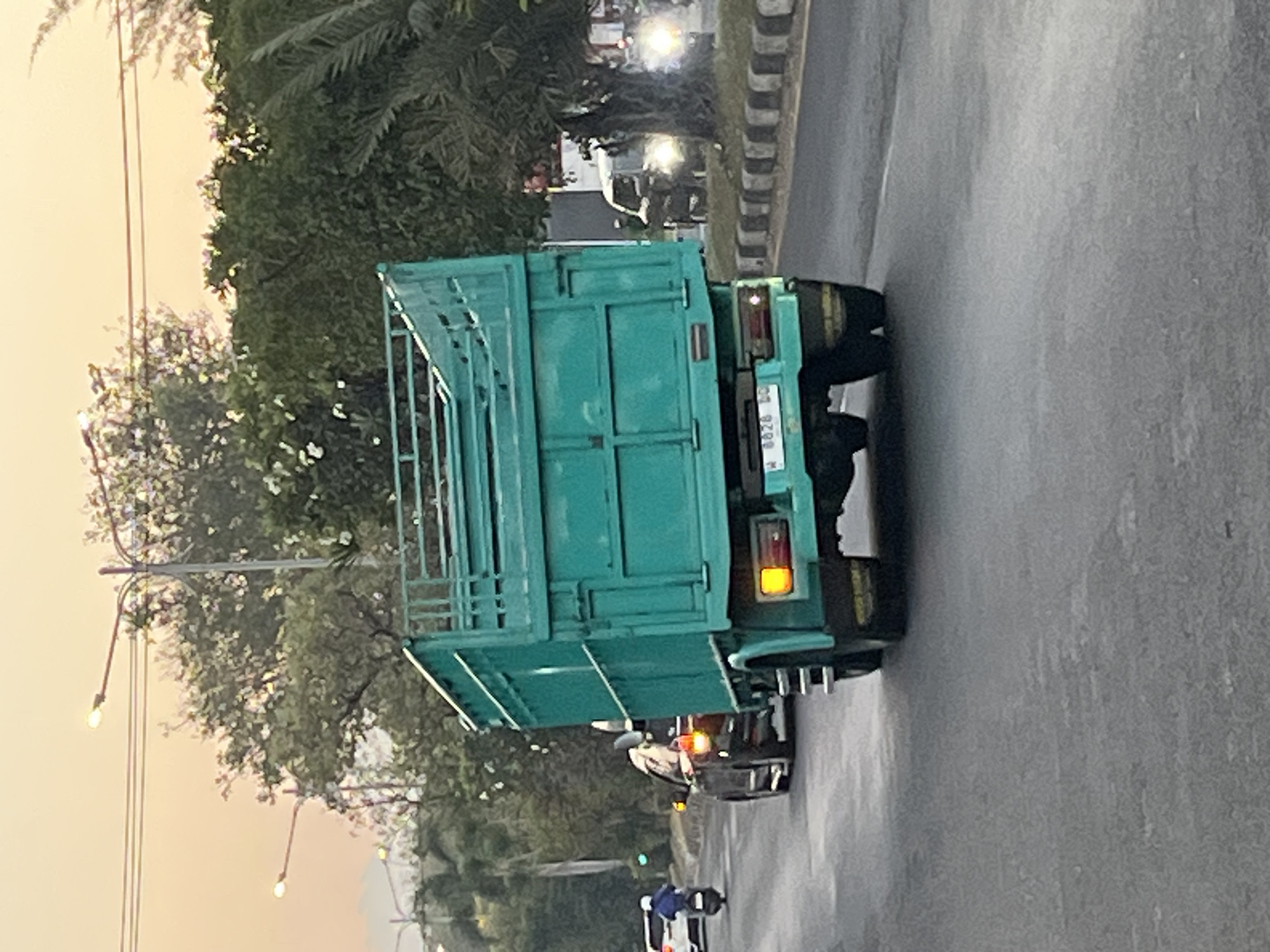 The image shows a teal colored truck driving on a road in Ngaban, Indonesia. The truck is in the foreground and takes up a significant portion of the frame. It has a large open cargo bed with metal bars around the top. The truck's license plate is visible and reads "W 6826 LQ".
In the background, other vehicles are present. To the left, a white car and a scooter with a rider are visible. To the right, a smaller white vehicle with its headlights on is driving. The environment appears to be a city street with trees and streetlights lining the road. The sky in the background is a soft orange and yellow, suggesting it is either sunrise or sunset. The road surface is asphalt and appears to be in decent condition. The scene is captured from a slightly low angle, looking up at the truck.