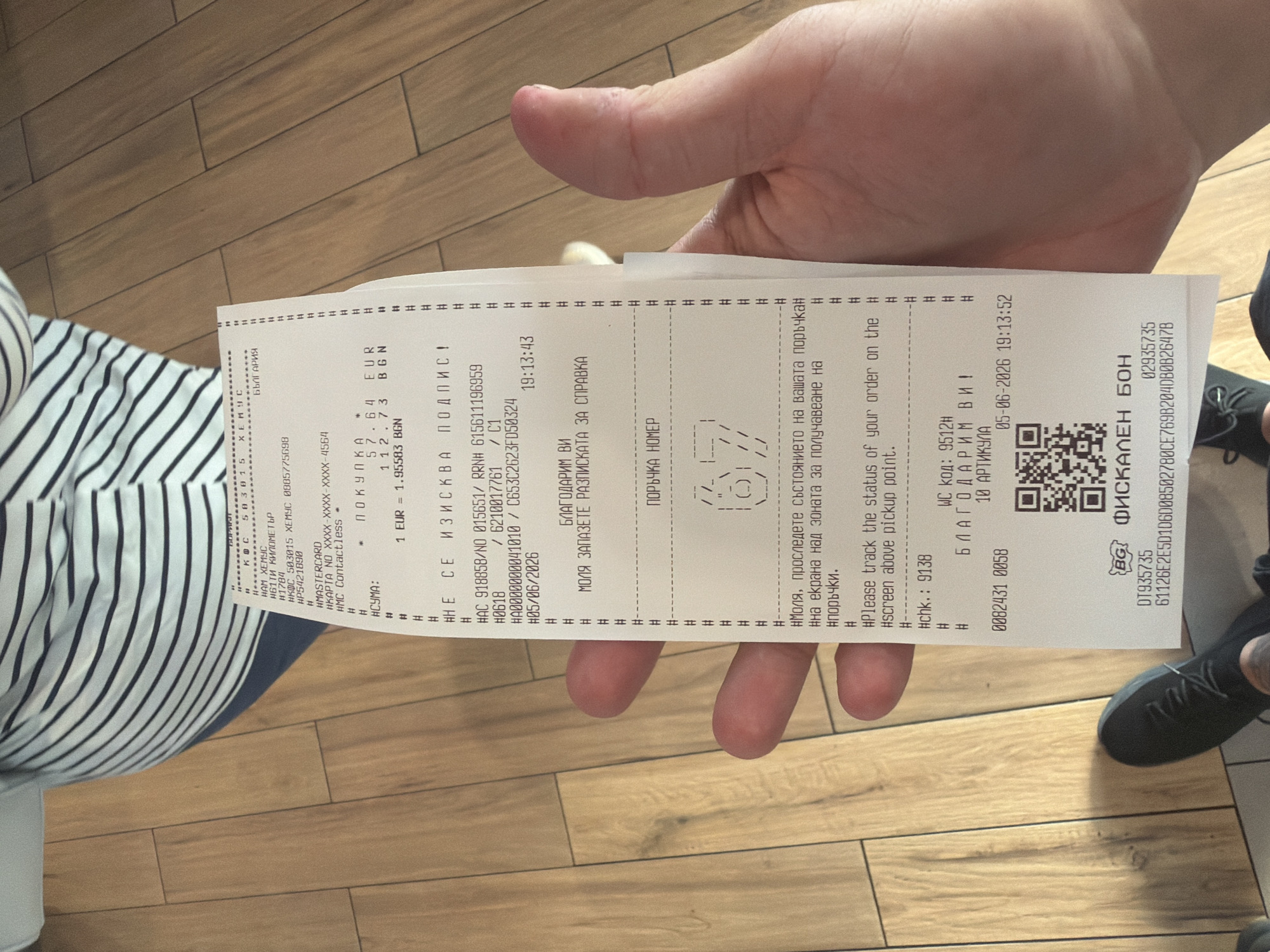 A paper receipt is held vertically in the left hand of an individual, positioned centrally in the image. The scene is an indoor environment, identified by the receipt as a KFC establishment ("КФС ХЕМУС") located near the Hemus Highway in Bulgaria, specifically in or around the city of Osikovitsa.
The individual's left hand, exhibiting discernible skin texture and visible knuckles, grips the receipt. Portions of the individual are visible in the background: above the receipt, a section of their torso is seen, clad in a horizontally striped shirt with alternating blue and white bands. Below the receipt, the individual's feet, shod in black sneakers, are resting on a light brown wooden floor composed of distinct planks.
The receipt details a transaction:
* **Establishment:** KFC, ID "КФС 50301", "КФС ХЕМУС" (KFC Hemus), Bulgaria.
* **Transaction:** A purchase totaling "57.64 EUR", equivalent to "112.73 BGN", with an exchange rate of "1 EUR = 1.95583 BGN". The payment was made via "НМАСТЕРСARD" (Mastercard), contactless, with the last four digits "4564" of the card number visible. No signature was required.
* **Identification:** Multiple transaction IDs are present, including "НАС 918858/NO 015651/ RRNH 615611196959" and other alphanumeric strings.
* **Date and Time:** The receipt displays a date of "05/06/2026" with times "19:13:43" and "19:13:52" at different points.
* **Order Information:** An order number "67" is depicted in ASCII art, and instructions are provided in both Bulgarian and English to "track the status of your order on the screen above pickup point."
* **Staff and Items:** "CHK.: 9138" and "WC код: 9512Н" are noted, and the purchase comprised "10 АРТИКУЛА" (10 items).
* **Fiscal Details:** The document is designated a "BG ФИСКАЛЕН БОН" (BG FISCAL RECEIPT) with unique identifiers "DT935735", "02935735", and a long fiscal code "G1126E2E5D1D6D0B50278CE76982D40BB2647B". A QR code is printed at the bottom, presumably linking to these fiscal details.